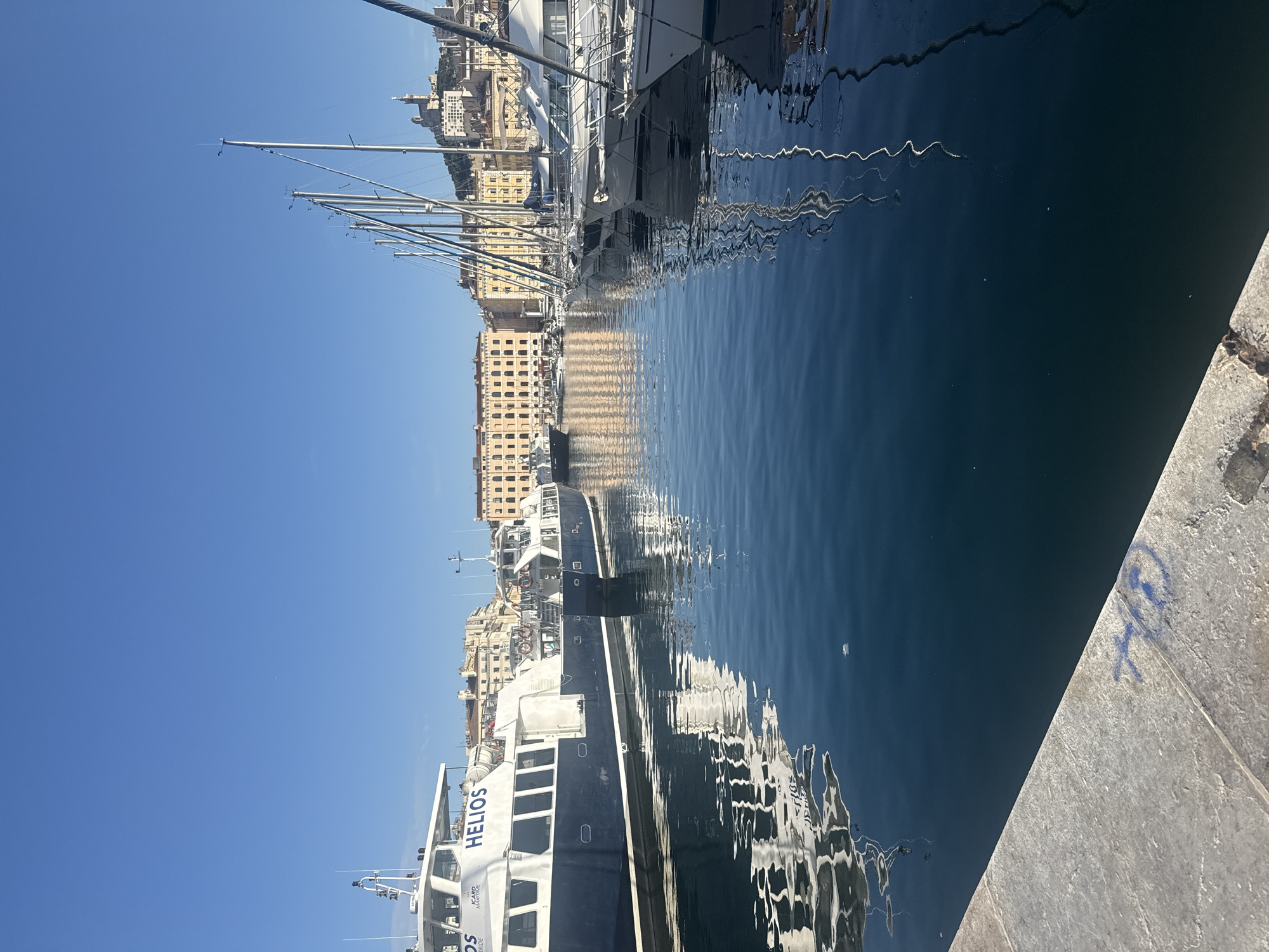 The image depicts a harbor scene under a clear blue sky. Several boats are docked in the water, with a prominent blue and white vessel on the left labeled "HELIOS." To the right, a line of sailboats with tall masts is visible. In the background, a cityscape of buildings with yellow facades and a distant hilltop with a structure resembling a church or basilica can be seen. The water is calm and reflects the boats and buildings. A concrete walkway with blue graffiti is in the foreground.