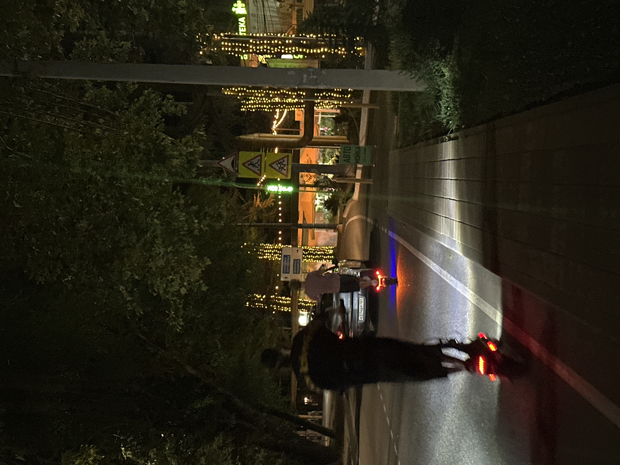 This is a nighttime shot of a street in the city of Sochi, Russia. The scene is illuminated by streetlights, decorative lights strung on trees and poles, and the lights from a car and two electric scooters. In the foreground, a person on a red-wheeled electric scooter is moving away from the camera. The scooter's taillights are bright red. Further down the street, another person on a similar scooter, with blue and orange lights, is moving away from the camera. A car is parked on the right side of the street, and its license plate is visible. The street is lined with trees and a sidewalk. A few street signs are also visible, including one with pedestrian symbols.