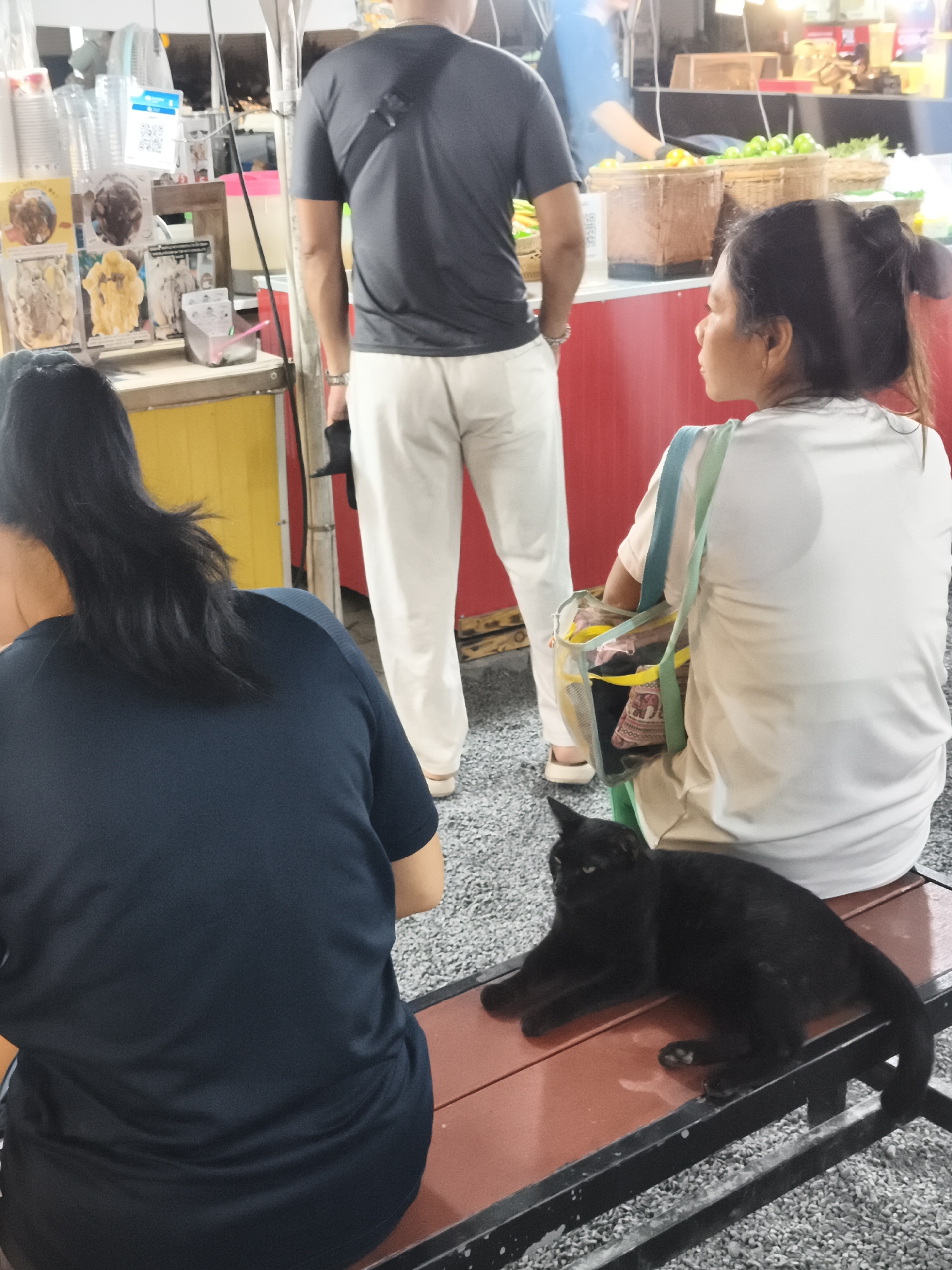 The image depicts an outdoor scene in บ้านบางรักษ์, Thailand. Two women are sitting on a red-brown bench with a black metal frame. A black cat is lying on the bench between them. A man wearing a dark gray t-shirt and white pants is standing in front of them with his back to the viewer. Behind him is a food vendor's stall with various items, including fruits and prepared food. The stall has a red and yellow color scheme. Other people are visible in the background. The ground is covered in small gravel.