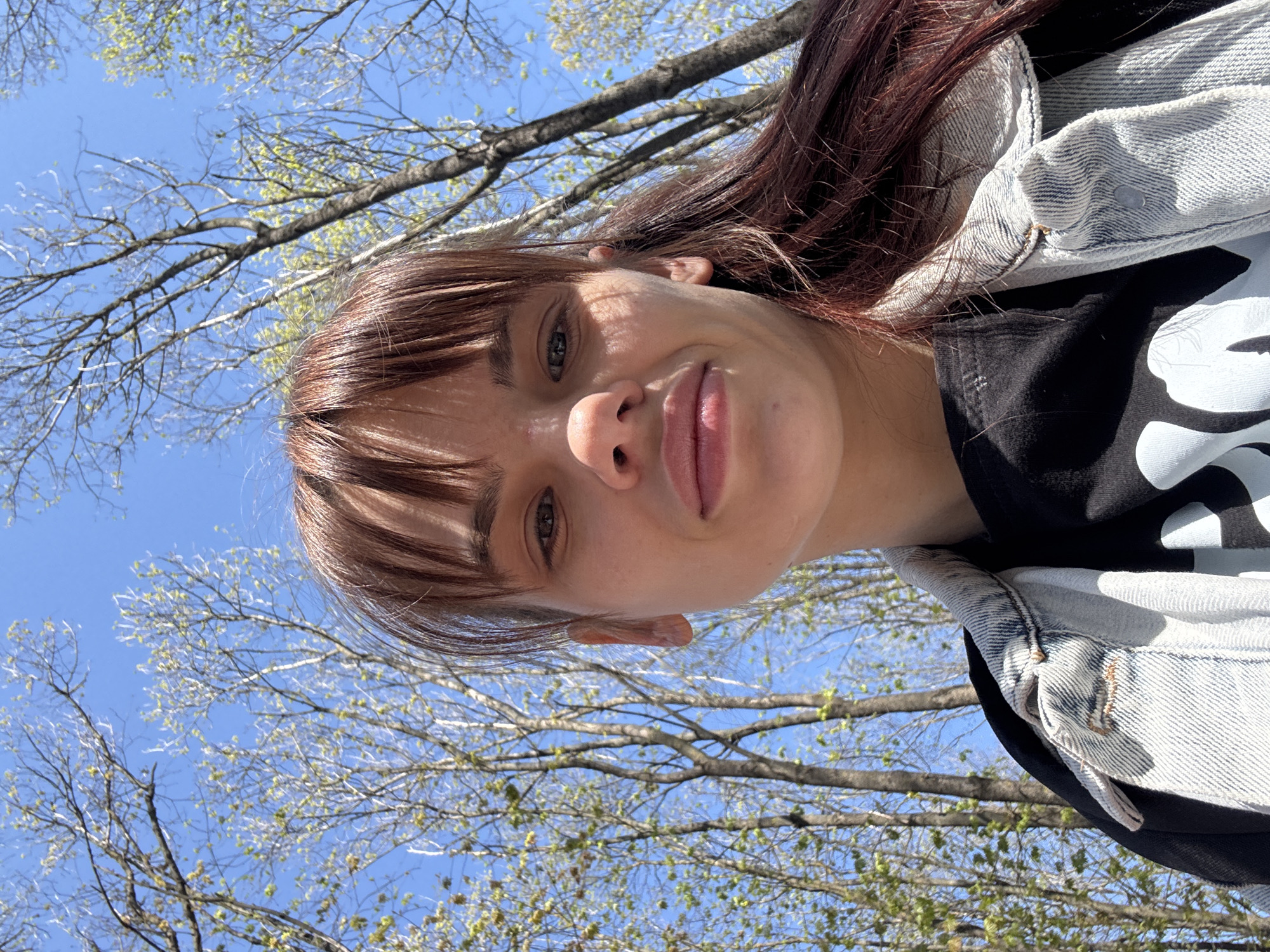 This outdoor selfie, taken in Rostov-on-Don, Russia, captures a young adult female from a low angle, looking upwards towards the camera. The subject's face is positioned on the left side of the frame, partially shaded by sunlight filtering through tree branches, creating a dappled light effect. She has medium-length brown hair with bangs, light-colored eyes (appearing green or hazel), and a subtle, relaxed expression with a slight smirk.
She is dressed in a light-wash denim jacket worn over a dark (likely black) t-shirt. A white graphic design, resembling the lower part of a stylized letter "S" or similar character, is visible on the t-shirt.
The background consists of a clear, bright blue sky dominating the upper and right portions of the image. Numerous tree branches, indicating early spring, are visible against the sky. These branches are just beginning to show small, light green leaves or buds, suggesting the seasonal transition. The overall impression is a sunny, pleasant day, likely mid-morning to late afternoon given the bright, direct sunlight. The upward perspective emphasizes the natural surroundings and open sky.