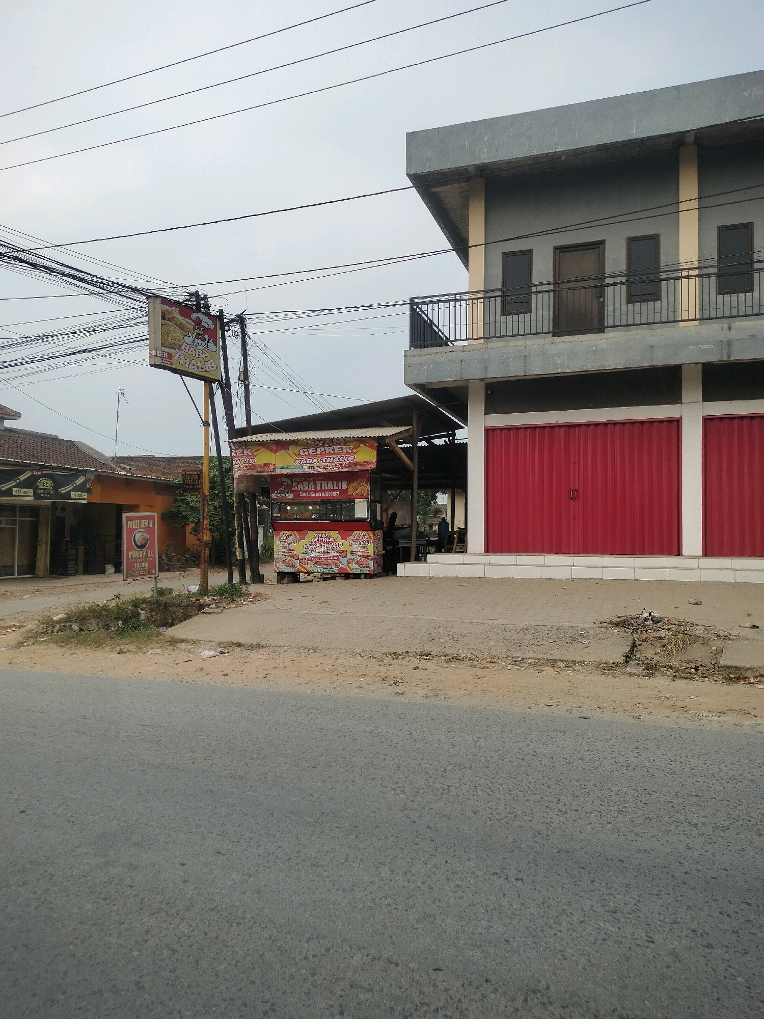 The image shows a street scene in Wayakroy, Indonesia, with a two-story building featuring gray walls and a balcony with a metal railing. The ground floor of the building has large red roller doors. Adjacent to the building is a food stall with a striped awning and red and yellow signage advertising "AYAM GEPREK CABA THALIB BABA THALIB". Several people are visible at the food stall, some appearing to be customers. In the foreground, a paved road runs alongside a dirt shoulder. Overhead, numerous electrical wires crisscross the sky, supported by poles. To the left of the food stall, another smaller building with an orange facade is visible, along with a sign that reads "PAKET HEMAT" (Hemat Package) and lists prices for "AYAM GEPREK". A yellow sign with an image of fried chicken and text is also prominently displayed on a pole near the food stall.