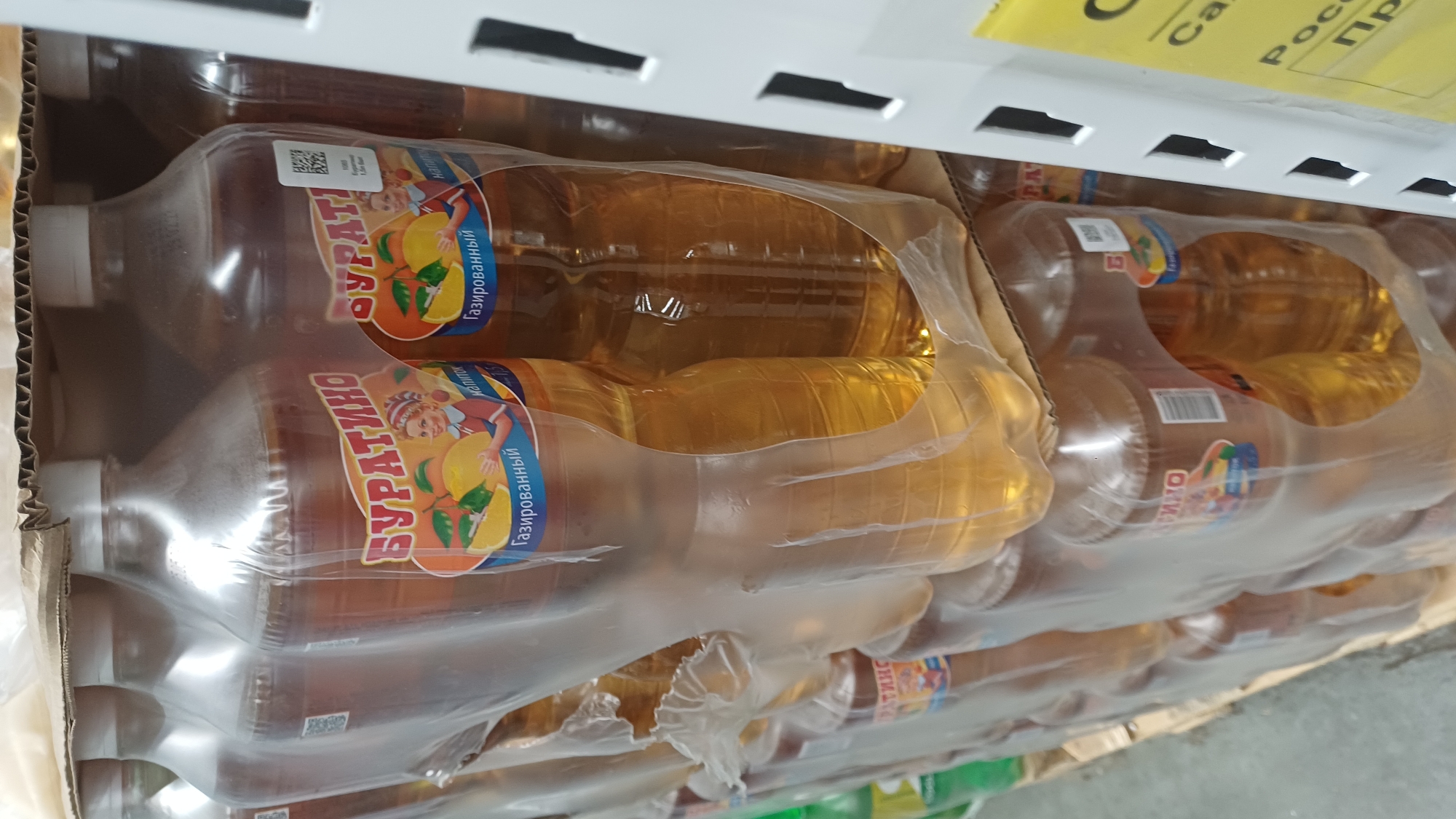 This is a close-up, eye-level, interior shot of a retail display shelf stocked with multiple plastic bottles of "Buratino" brand carbonated beverage. The bottles are clear plastic and contain a golden-yellow liquid. They are shrink-wrapped together in packs. The labels on the bottles feature a cartoon illustration of a young girl with blonde hair holding a lemon, with the text "Буратино" (Buratino) and "Газированный напиток" (carbonated drink). The bottles are stacked in two rows, with the top row partially visible. The shelving unit is white and appears to be metal. A price tag or promotional sign in yellow with Russian text is partially visible on the right side of the frame. The background is blurred but suggests a retail environment. No people are visible in the image. The scene is indoors, likely a supermarket or convenience store in Izhevsk, Russia, given the Cyrillic lettering.