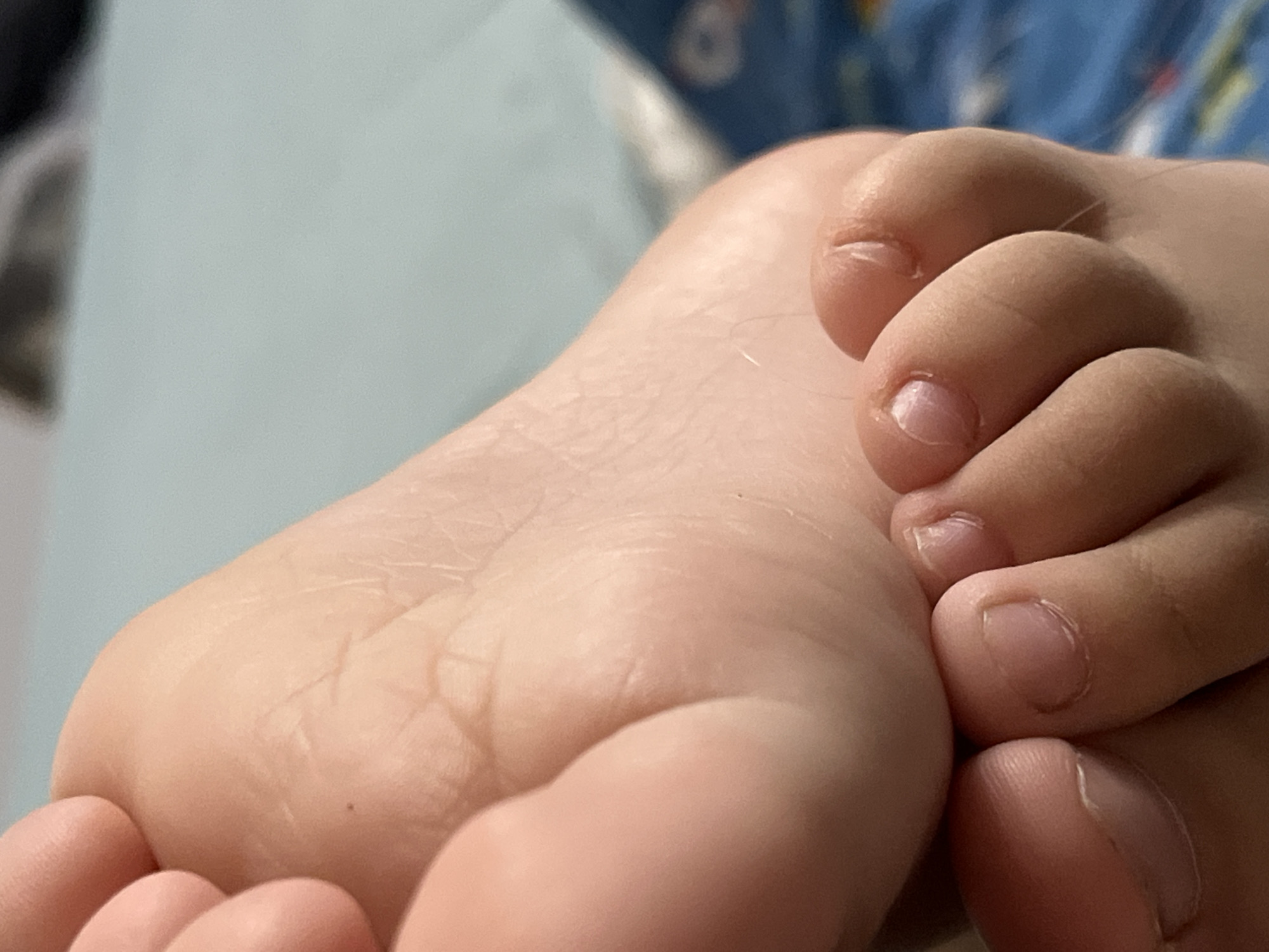A close-up, eye-level shot focuses on the sole and toes of a child's foot. The skin is pale and appears soft, with visible creases on the sole and subtle lines around the toe joints. The toenails are short and neat. One toe is gently pressed against the sole. The background consists of a soft, pale blue surface, likely bedding, and a glimpse of a patterned blue fabric. The lighting is soft and diffused, illuminating the details of the foot without harsh shadows. The image does not depict any identifiable elements of the city of Krasnyye Barrikady, Russia, or any specific setting within a city.