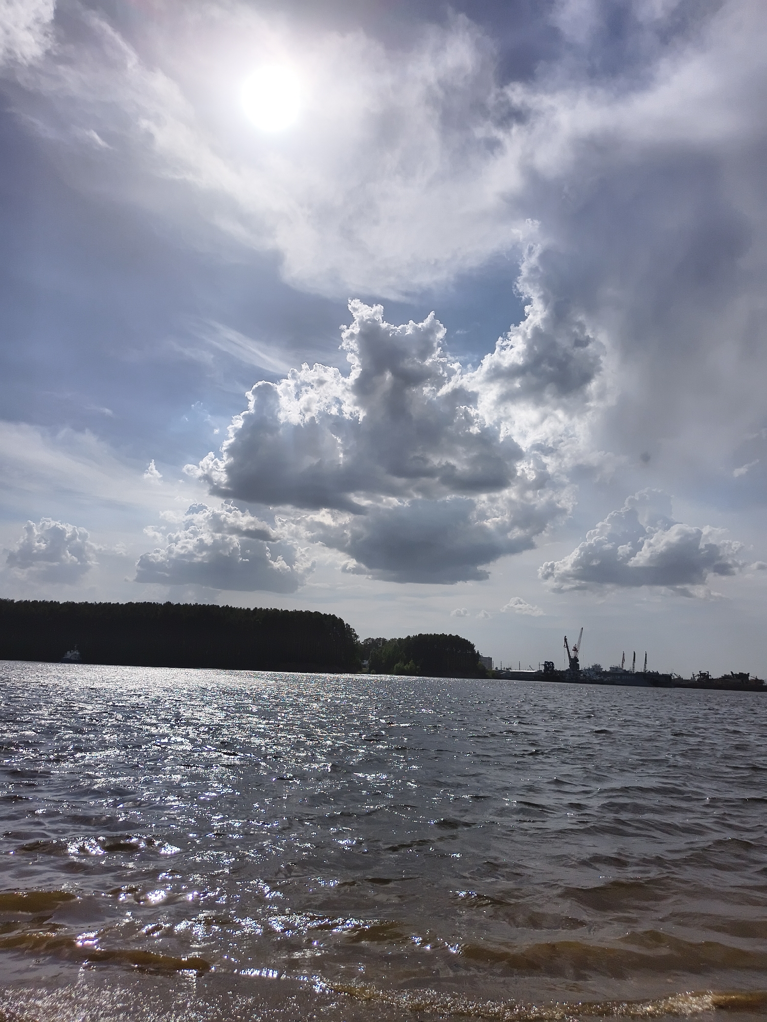 The image shows a body of water under a cloudy sky with the sun visible. A dark treeline is visible on the opposite shore, with cranes and industrial structures further in the distance. The water's surface is rippled, reflecting light. The location is in the city of Unknown, Unknown.