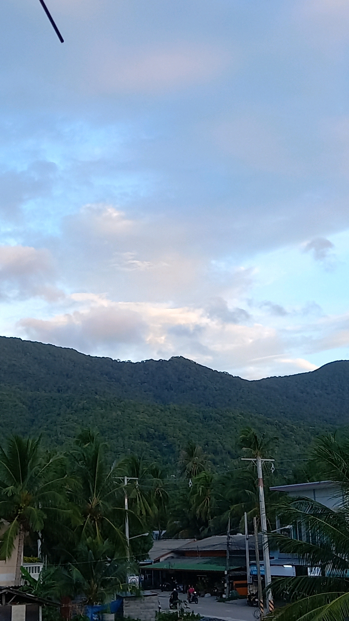 This is a vertical, medium-range, eye-level shot of a town on Ko Pha-ngan District, Thailand, at dusk. In the foreground, lush green palm trees frame the scene. A road runs through the middle ground, with several buildings and shops lining its sides. A few motorcycles are parked and being ridden on the road. In the background, a dense, tree-covered mountain range dominates the horizon. The sky above is a pale blue with scattered, wispy clouds, tinged with the soft light of twilight. A thin, dark, linear object extends from the top left corner of the frame, partially obscuring the sky.