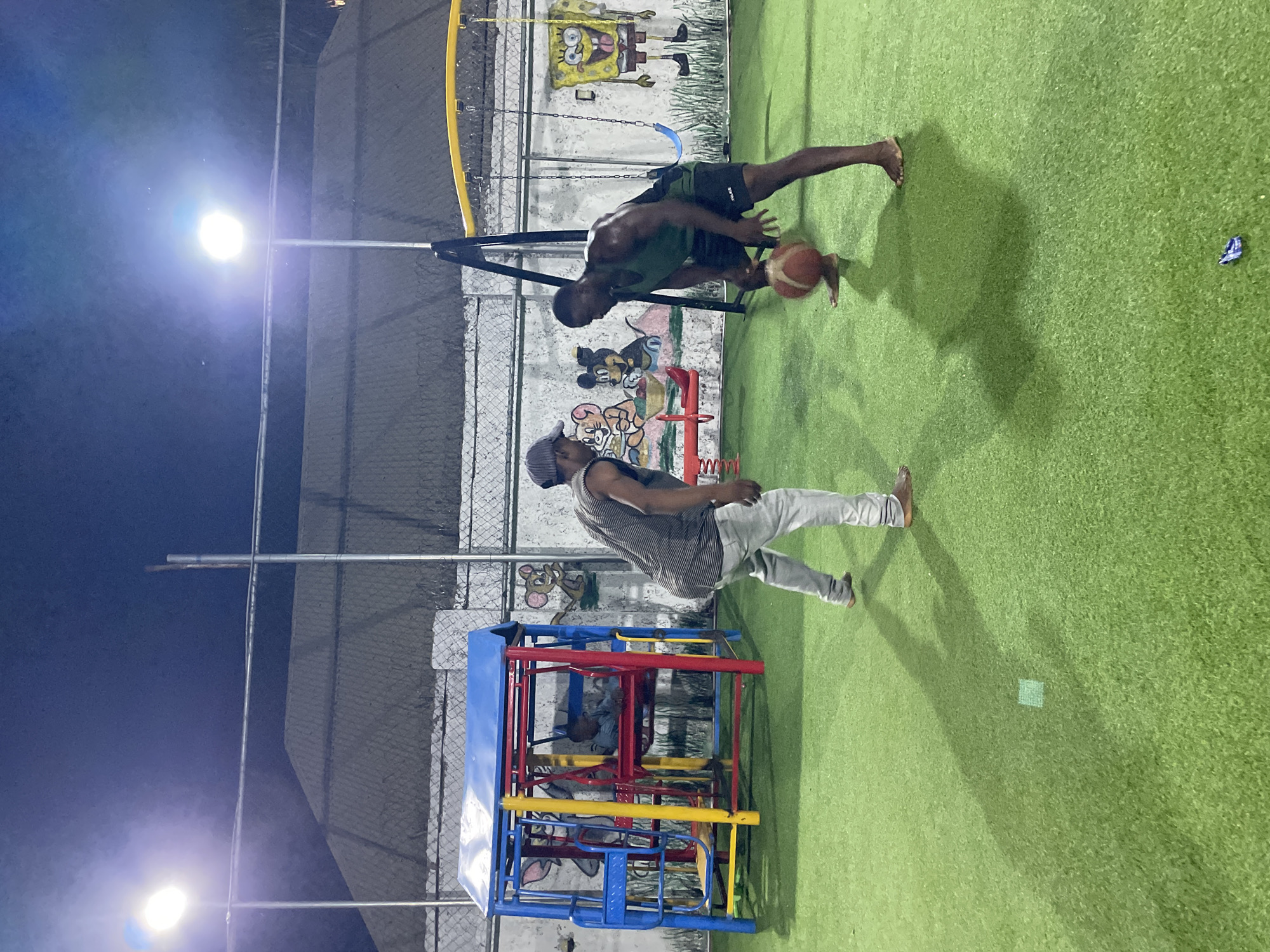 Two dark-skinned men are on an artificial turf sports field at night. The man closest to the foreground wears a gray cap, a striped tank top, gray pants, and no shoes. He is walking. The other man is darker-skinned, wearing a green tank top and dark shorts. He is dribbling a basketball. Behind them is a muraled wall and a playground structure. Lampposts illuminate the scene. The location is likely Akwanga, Nigeria.