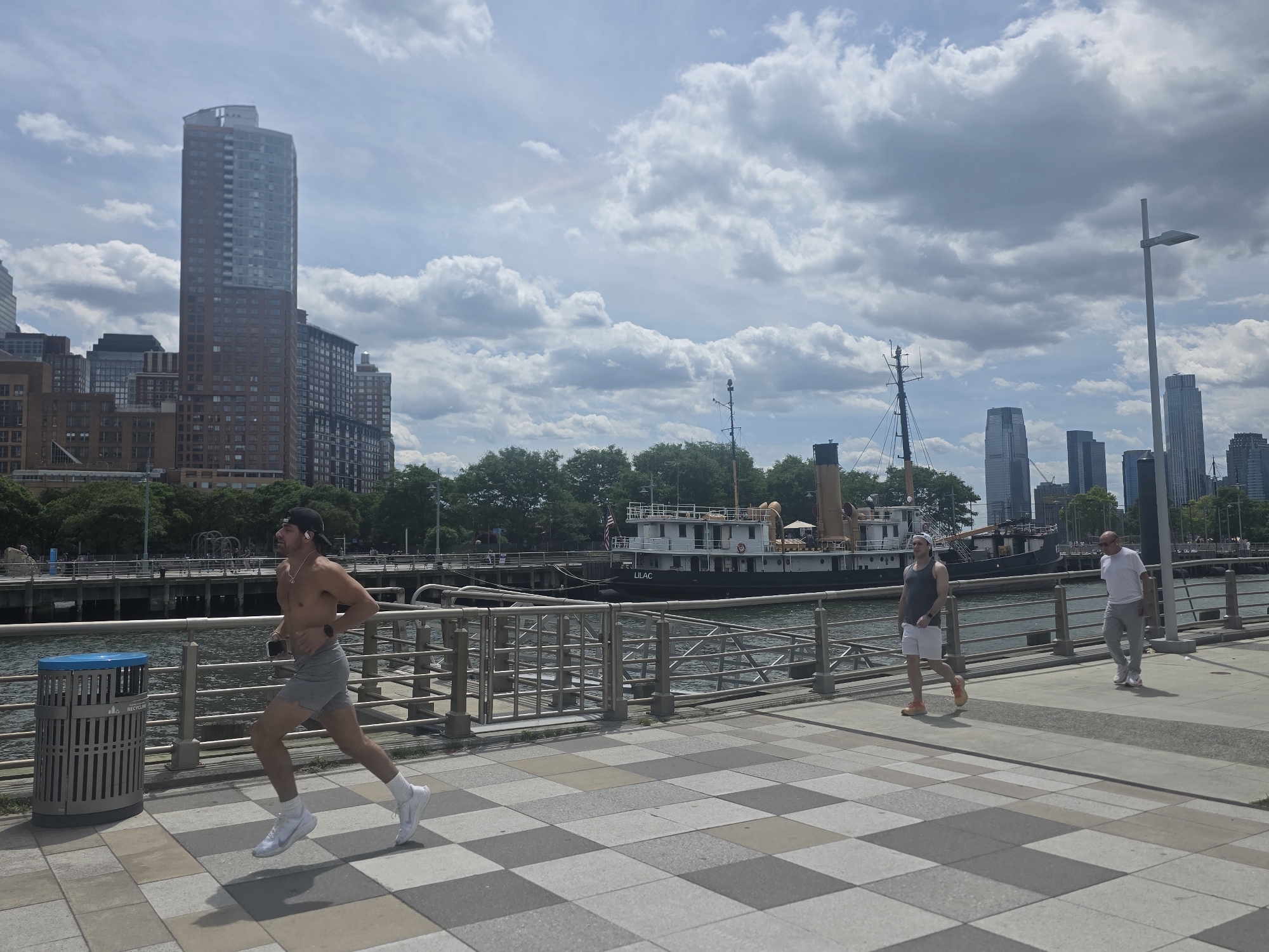 The image shows a waterfront area in New York City, United States of America. In the foreground, a shirtless man is running on a gray and white tiled walkway. Further back, two men are walking. A docked ship is visible behind the walkway, and in the background, there are numerous buildings and skyscrapers. The sky is overcast with heavy cloud cover. A light pole and metal fencing are also present along the walkway.