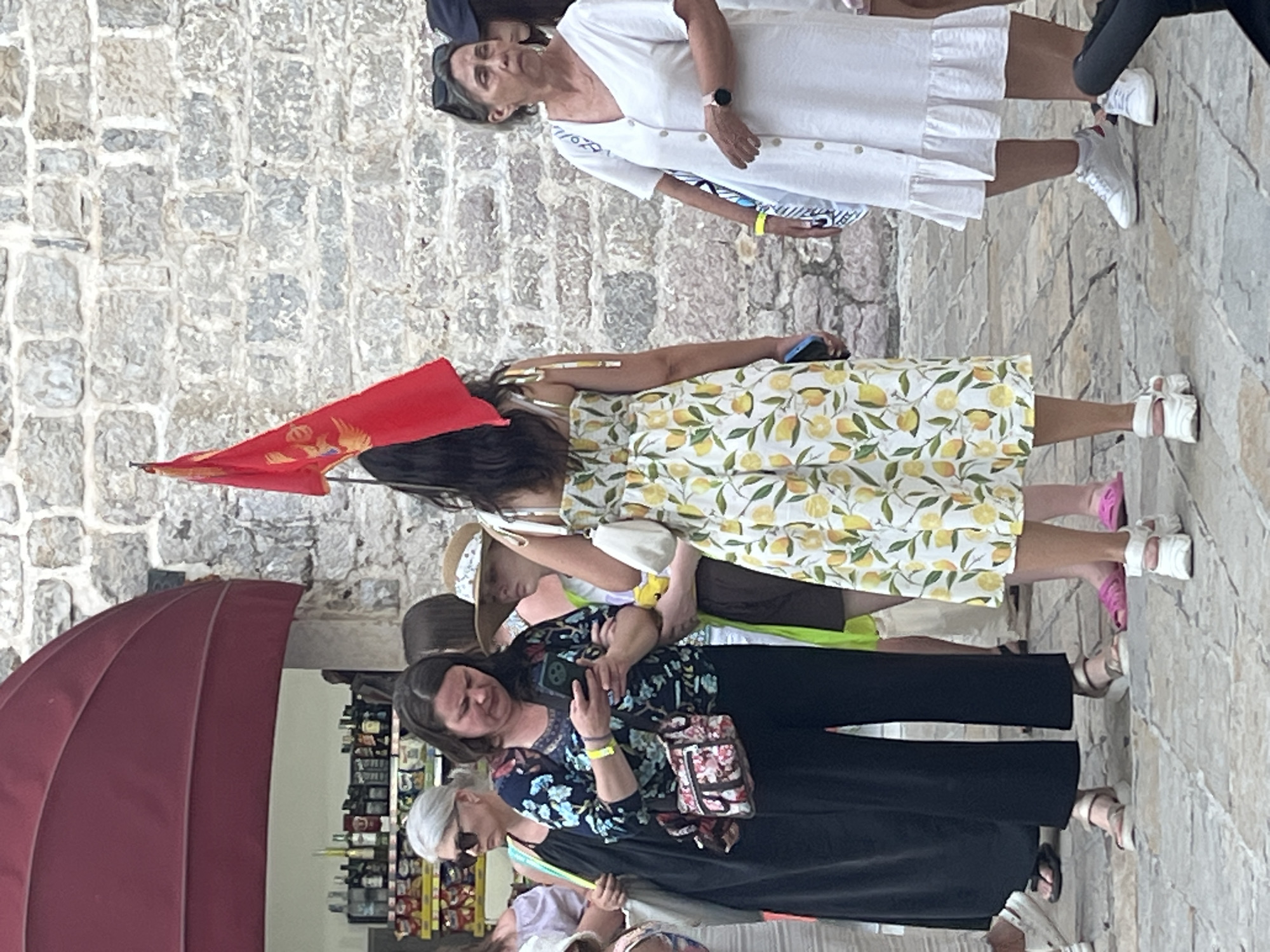 The image depicts a street scene in Budva, Montenegro, possibly near the old town. Several individuals are present, most of whom are women. In the foreground, a woman wearing a yellow dress with a lemon pattern is holding a smartphone. To her left, another woman in a black dress is looking at her phone. A woman with gray hair and sunglasses stands further to the left. In the background, a red flag with a golden emblem, likely the flag of Montenegro, is visible. The environment appears to be an outdoor area with stone buildings and a stall selling various items, including bottles of alcohol and snacks. The ground is paved with cobblestones. The overall atmosphere suggests a casual outing or a tourist gathering.