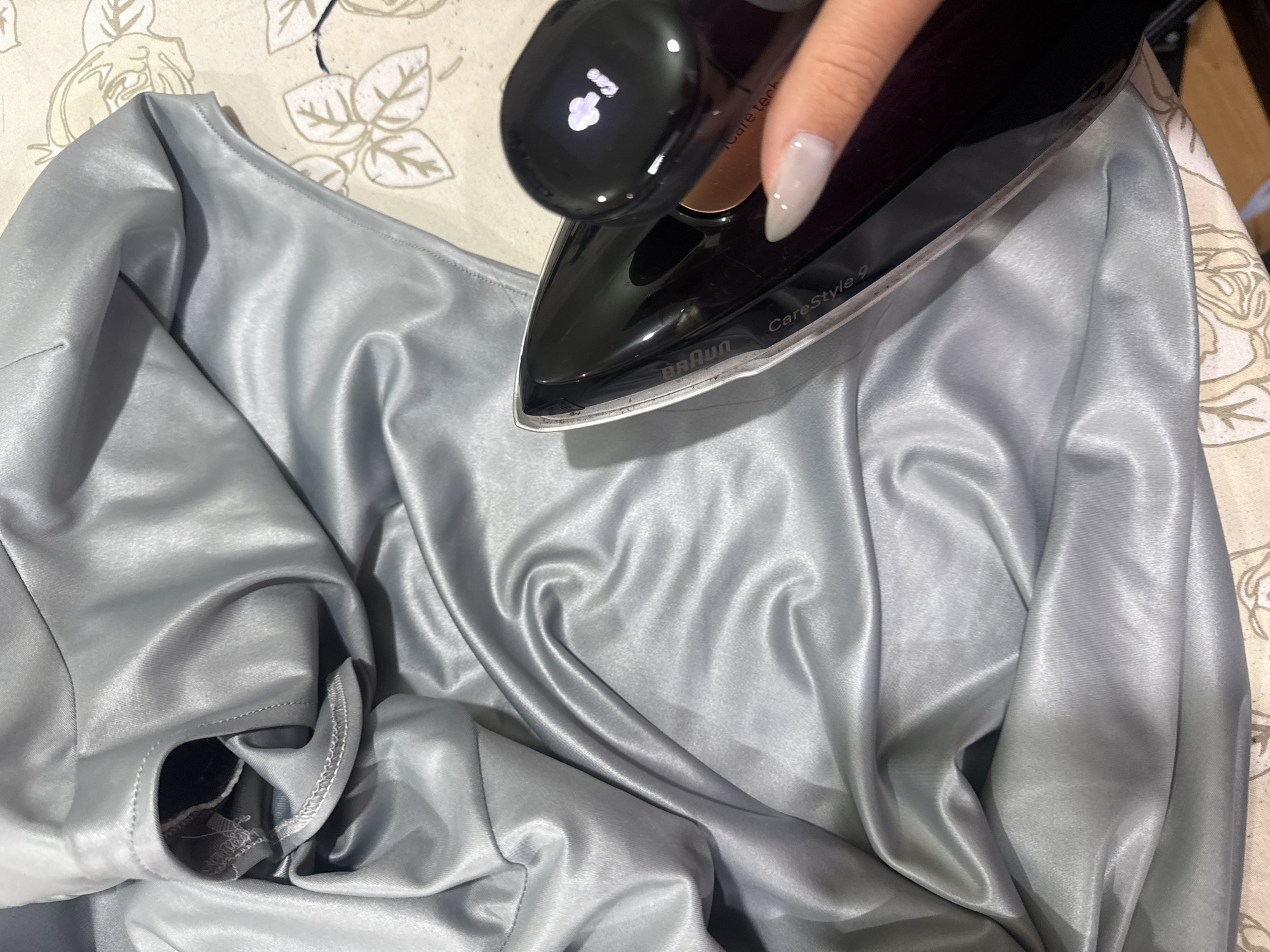 An overhead shot captures a close-up scene of ironing taking place in Yerevan, Armenia. The primary subjects are a person's hand, a black Braun iron, and a piece of grey or silver satin-like fabric.
The person's hand, with neatly manicured, light-colored polished nails, is guiding the iron over the fabric. The iron, specifically a "Braun CareStyle 9" model, is positioned at the center, its black body and silver soleplate contrasting with the material below. The iron's digital display shows "iCare tech" along with what appears to be a temperature or setting icon. Some residue or wear is visible along the edge of the iron's soleplate.
The fabric itself is a smooth, reflective material, likely satin or a similar synthetic, exhibiting a lustrous sheen. It is draped and gathered, showing some wrinkles that the ironing process is intended to smooth out. The fabric has visible stitching along some edges and seams, suggesting it is part of a garment or textile item.
The ironing is occurring on an ironing board, partially visible beneath the fabric and iron. The ironing board cover features a light-colored, subtle floral or leafy pattern against a cream background. The overall setting appears to be indoors, likely within a home. No specific time of day or emotions are discernible from the image.