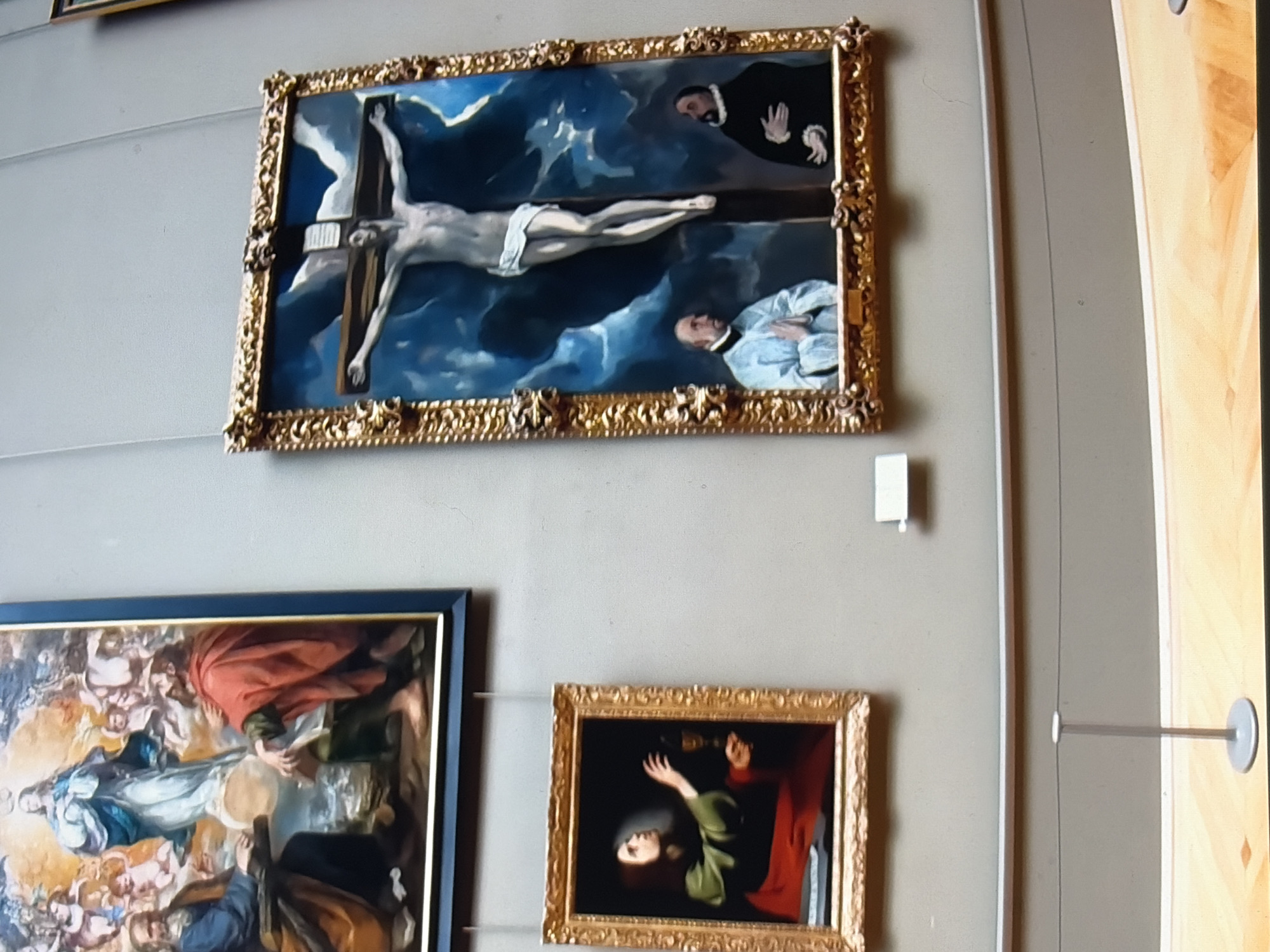 The image displays a section of an art gallery with three paintings hanging on a light gray wall. In the upper left, a large framed painting depicts a religious scene with multiple figures, including a central figure in blue and white, surrounded by other individuals and cherubs. To the right, a tall, narrow framed painting shows a crucifix with Jesus Christ, rendered with a pale, ethereal body against a dramatic blue sky with clouds. Below the crucifix, two figures in dark attire are visible. In the lower left, a smaller, square framed painting features a figure in a green robe and red drapery, holding a chalice and looking upwards. A thin metal stanchion with a wire barrier is positioned in the foreground, separating the viewer from the artwork. The floor is a light wood.