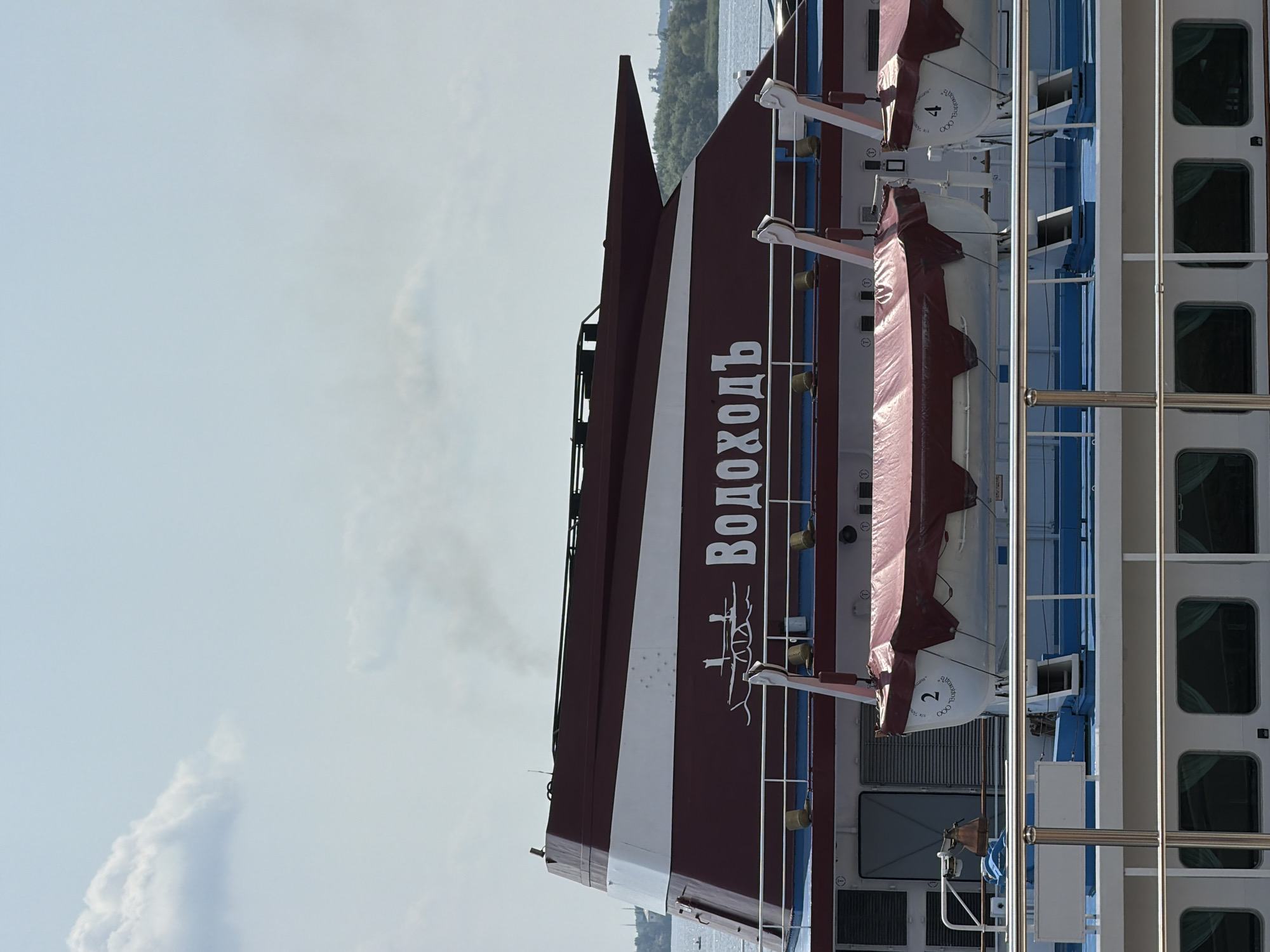 The image shows a close-up view of the upper deck of a river cruise ship docked in Saratov, Russia. The ship has a distinctive maroon and white striped superstructure. The name "Водоходъ" (Vodokhod), written in white Cyrillic script, is prominently displayed on the maroon section of the superstructure. Below the superstructure, two lifeboats are visible, covered with maroon tarpaulins, each with a number (2 and 4) and some text in Cyrillic on the side. The lifeboats are mounted on davits. The ship's railing, painted white and silver, runs along the deck. The blue deck surface is visible between the lifeboats. In the background, there is a body of water, trees on the distant shore, and a cloudy sky. The overall impression is of a serene maritime scene in an urban environment.