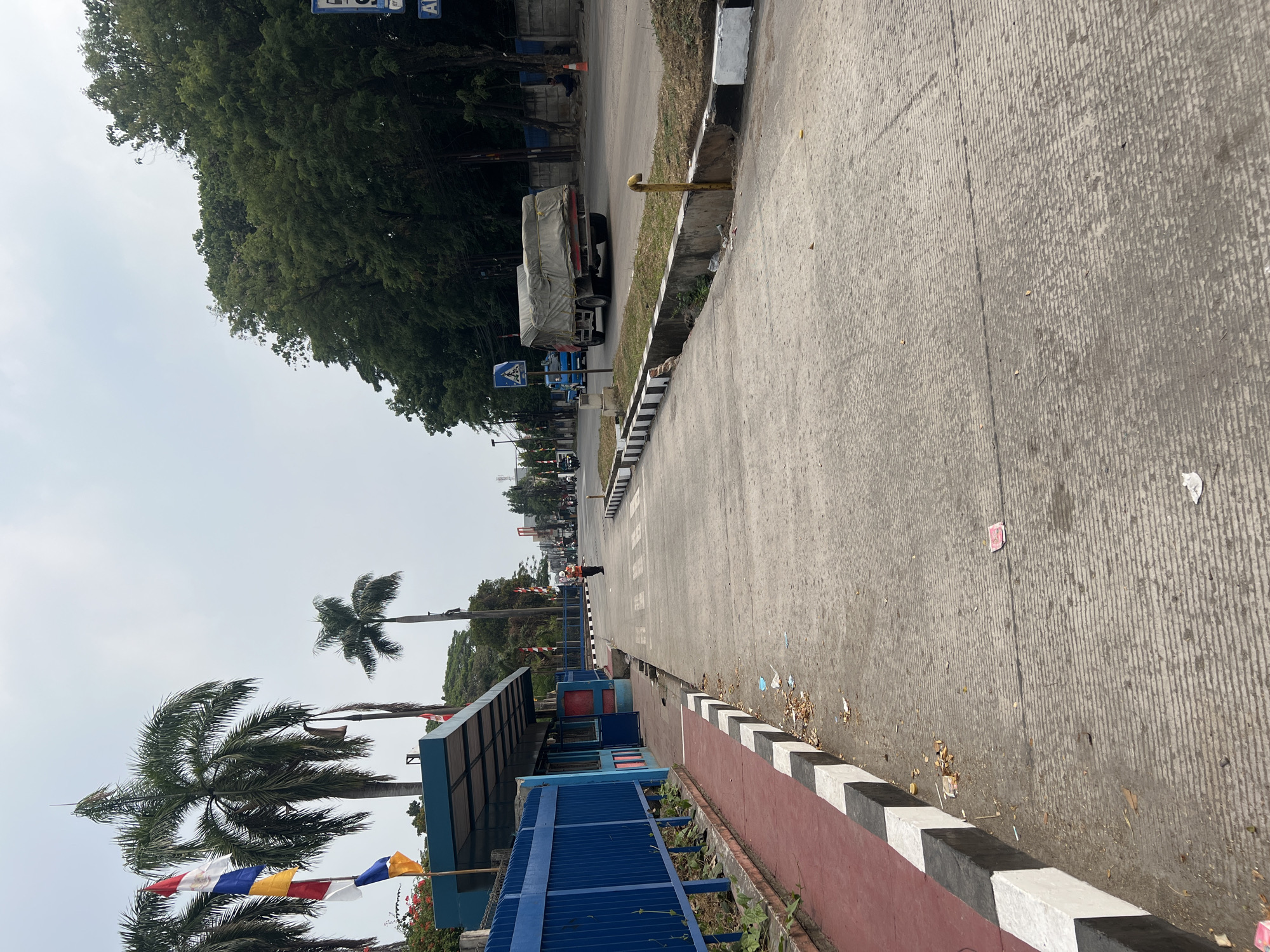 The image shows a street scene in Citeureup, Indonesia, with a paved road and sidewalk. On the left side of the image, there is a blue fence and a covered structure with a blue roof, possibly a bus stop or waiting area. Several flags are visible on poles near the structure. A pedestrian crossing is marked on the road. Further down the road, a truck covered with a tarp is driving, and another blue truck is visible in the background. Trees line the right side of the road. The sky is overcast. There are a few people visible on the street, including one person wearing an orange vest. Debris is scattered on the sidewalk.