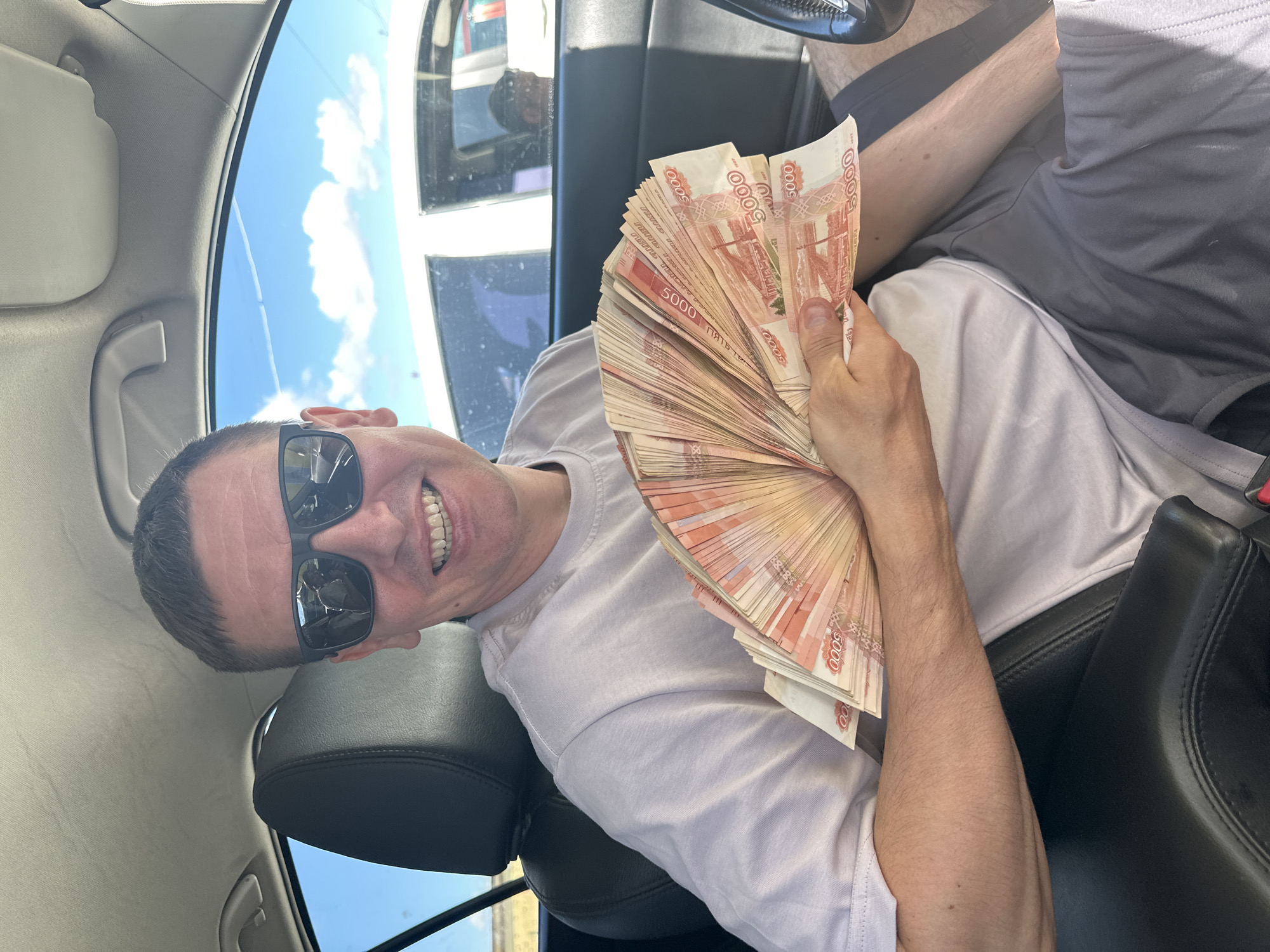 A man with short dark hair and sunglasses is sitting in the driver's seat of a car. He is holding a large fan of 5000 ruble banknotes in his left hand. The man is wearing a light-colored t-shirt and gray shorts. The interior of the car is visible, including the black leather seat and steering wheel. The background shows a bright blue sky with some clouds and another vehicle. It is daytime, and the lighting suggests it is sunny. The man is smiling and looking towards the camera. There is no specific indication of the location within Chaikovsky, Russia.