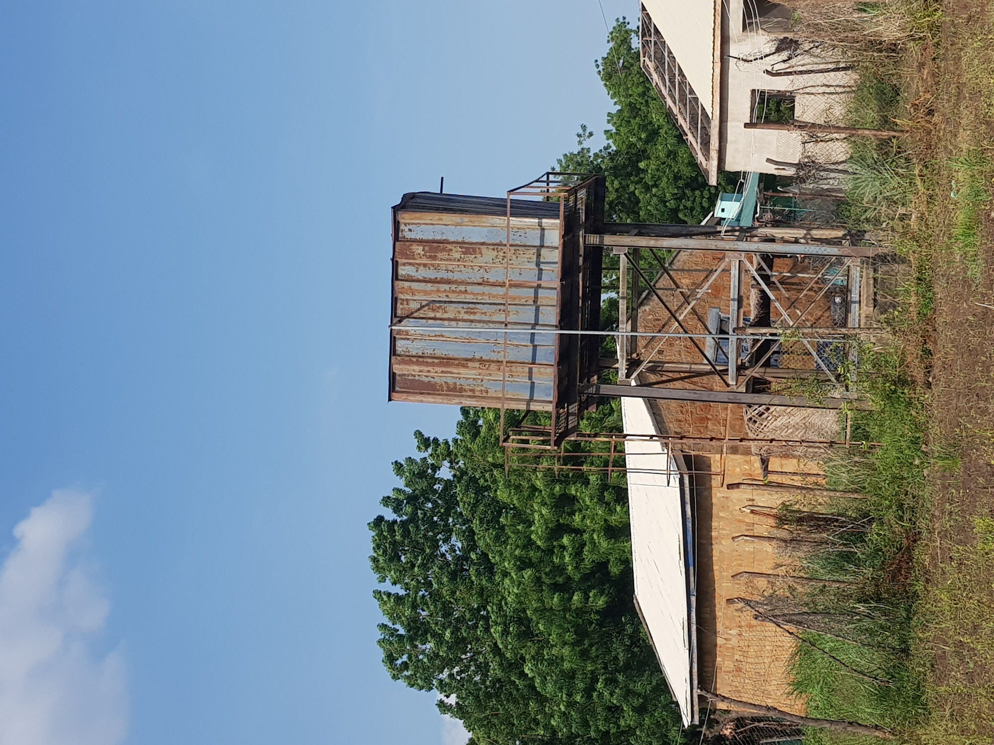 This image shows a rusty, elevated water tank situated in Jimeta, Nigeria, under a clear blue sky. The tank is supported by a metal structure. To the left of the water tank is a building with a light-colored roof and brick walls. In front of the building is a fence made of wire mesh and wooden posts, with overgrown vegetation. To the right of the water tank is another building, partially constructed or in disrepair, with a light-colored roof and visible roof beams. Dense green trees are visible behind the water tank and buildings. The ground in the foreground is covered with dry, brown vegetation and soil. There are no people visible in the image.
