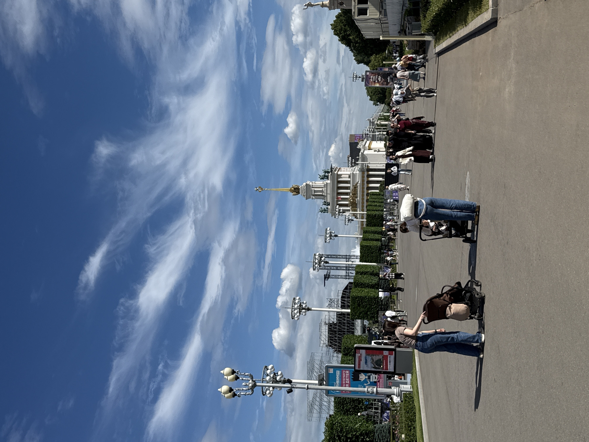 This is a vertical, eye-level, outdoor, medium shot taken on a sunny day in Moscow, Russia, at the VDNKh exhibition center. In the foreground, a person wearing blue jeans and a brown shirt bends over a stroller, while another person in similar attire stands nearby. The asphalt pathway stretches into the distance, populated by numerous people walking in both directions.
In the mid-ground, iconic architecture of VDNKh is visible, including a grand white pavilion topped with a golden spire and stars, flanked by other ornate structures. Decorative lampposts and neatly trimmed hedges line the pathway. In the background, a bright blue sky is adorned with wispy white clouds and dynamic streaks of cirrus clouds. Further back, scaffolding and other temporary structures are present, suggesting ongoing events or construction. The overall atmosphere is lively and suggests a public gathering or event at a renowned cultural landmark.