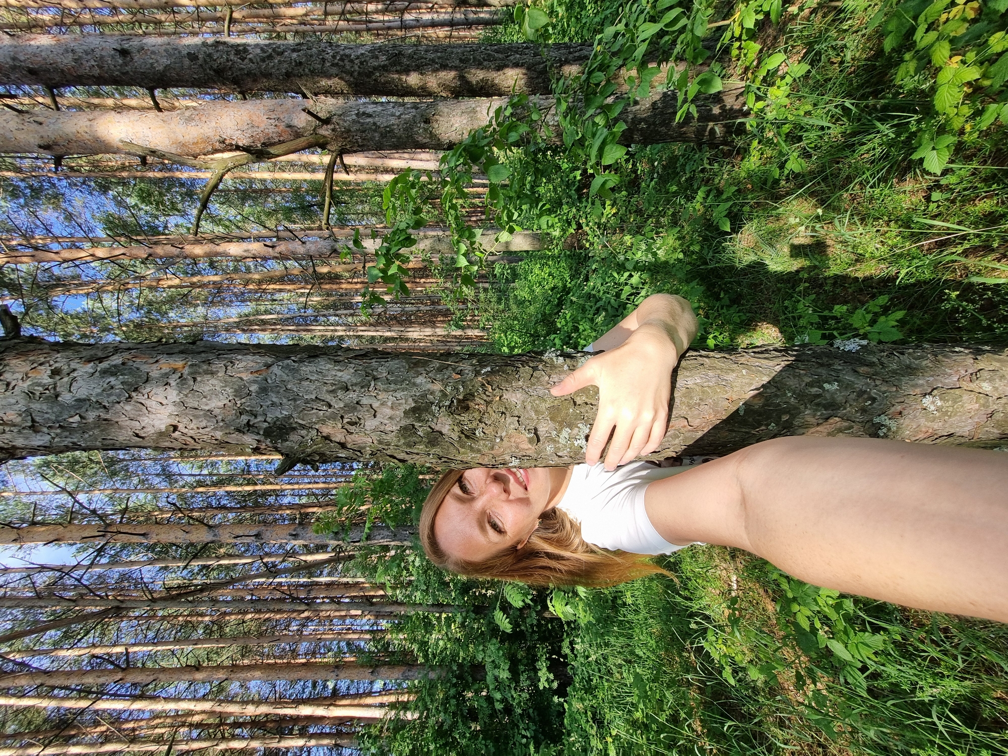 The image shows a woman in a forest, with pine trees and lush green foliage. The woman is lying on her side, with her head resting on a tree trunk. She is smiling and looking at the camera. Her left hand is reaching out and touching the tree trunk. She is wearing a white t-shirt and white socks. The sun is shining through the trees, casting dappled shadows on the forest floor. The image appears to have been taken on a clear day, possibly in the late afternoon, given the warm lighting. The natural setting suggests a recreational activity, such as resting or enjoying nature. There is no visible text in the image.