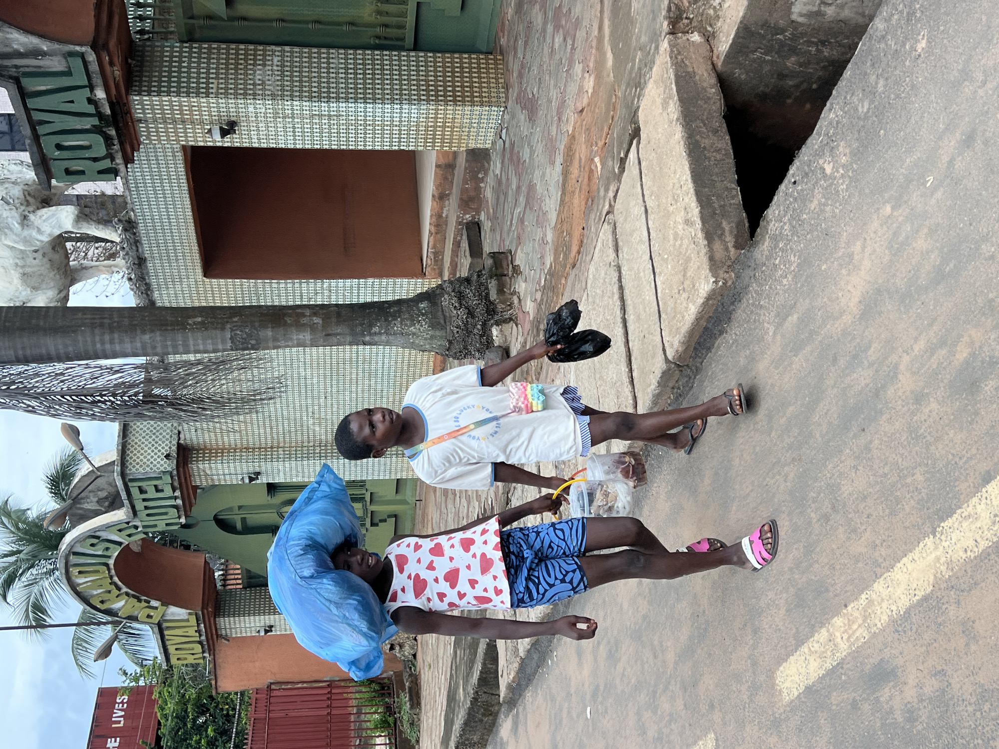 Two children walk on a paved road in Nkpor, Nigeria, in front of a building with "ROYAL PARADISE HOTEL" signage. The child on the left, a girl, carries a large blue plastic bag on her head and wears a white tank top with red hearts and blue shorts. She also carries a clear plastic bucket in her right hand, containing some items. The child on the right, a boy, wears a white t-shirt with text and blue and white striped shorts, and a small colorful purse hangs across his body. He carries two black plastic bags in his left hand. The environment includes a sidewalk made of concrete slabs, a drainage ditch, and palm trees. The building facade is decorated with patterned tiles and features an archway. A statue of a white animal, possibly a horse, is visible on the roof of the building. The sky is overcast.