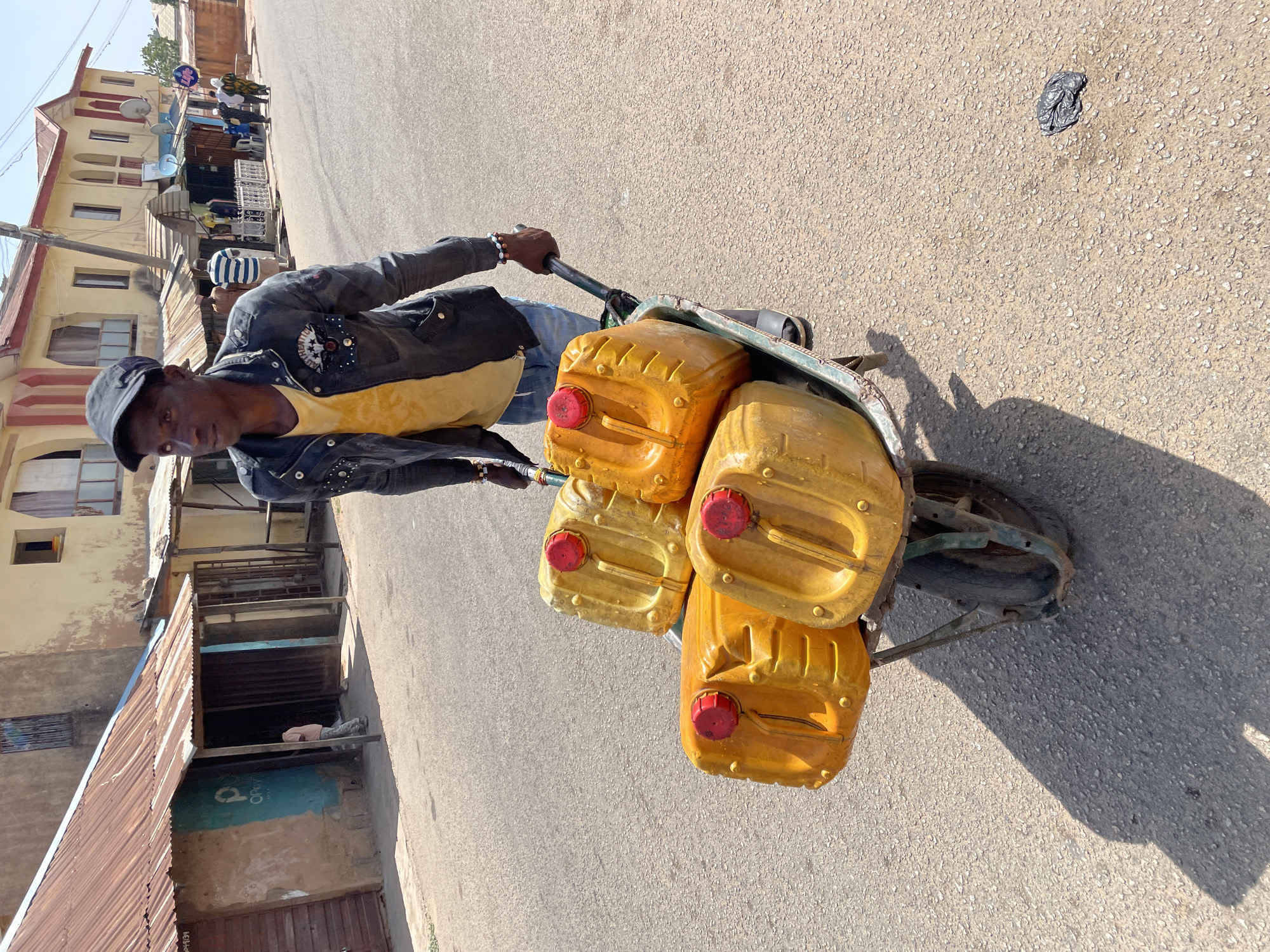 In a street in Jos, Nigeria, a man is pulling a green wheelbarrow loaded with four yellow, plastic jerrycans with red caps. He wears a dark cap, yellow shirt, jeans, and a black jacket. The street is paved with asphalt. Buildings with corrugated metal roofs and multi-story structures are visible in the background. A piece of dark debris lies on the road.