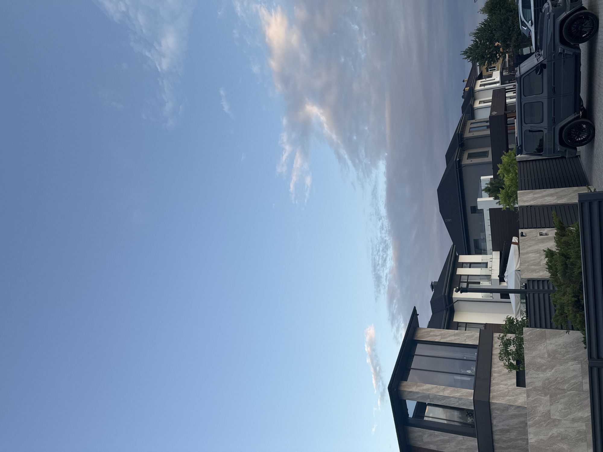 The image depicts a row of modern, multi-story houses under a clear blue sky with scattered clouds. The architecture is contemporary, featuring large windows, geometric shapes, and a mix of grey stone and dark materials. Several houses are visible, suggesting a residential neighborhood.
In the foreground, a dark grey, rugged-looking SUV, possibly a Mercedes-Benz G-Class, is parked on a paved driveway. The vehicle's tinted windows and black wheels contribute to its imposing presence. Adjacent to the houses is a dark metal fence. Lush green trees and shrubs are interspersed around the properties, adding a touch of nature to the urban setting.
The lighting and cloud formations suggest it is either early morning or late afternoon. The sky is bright, with some wispy clouds tinged with pale pink and orange, indicating the sun is low on the horizon. The overall mood is serene and upscale.
There is no visible text in the image. The context provided indicates the location as Yekaterinburg, Russia.