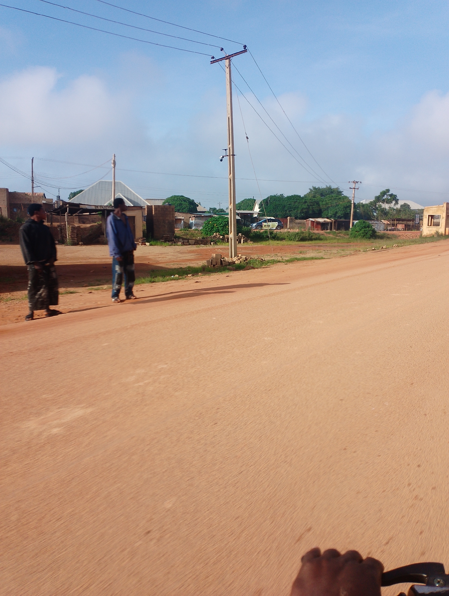 The image depicts a scene in Kwang, Nigeria, featuring a dusty, reddish-brown dirt road in the foreground. On the left side of the road, two young men are standing. The man closer to the camera is wearing a black long-sleeved shirt and patterned trousers. The man further back is wearing a blue hooded jacket and dark trousers. In the background, there are several small buildings with corrugated metal roofs and brick or mud walls. Some are partially constructed or in a state of disrepair. There are also trees and vegetation lining the background. Utility poles with overhead wires are visible throughout the scene, indicating the presence of electricity. A white vehicle with writing on its side is parked in the middle ground. The sky is partly cloudy with a clear blue sky dominating. The overall impression is of a rural or semi-urban environment in Nigeria.