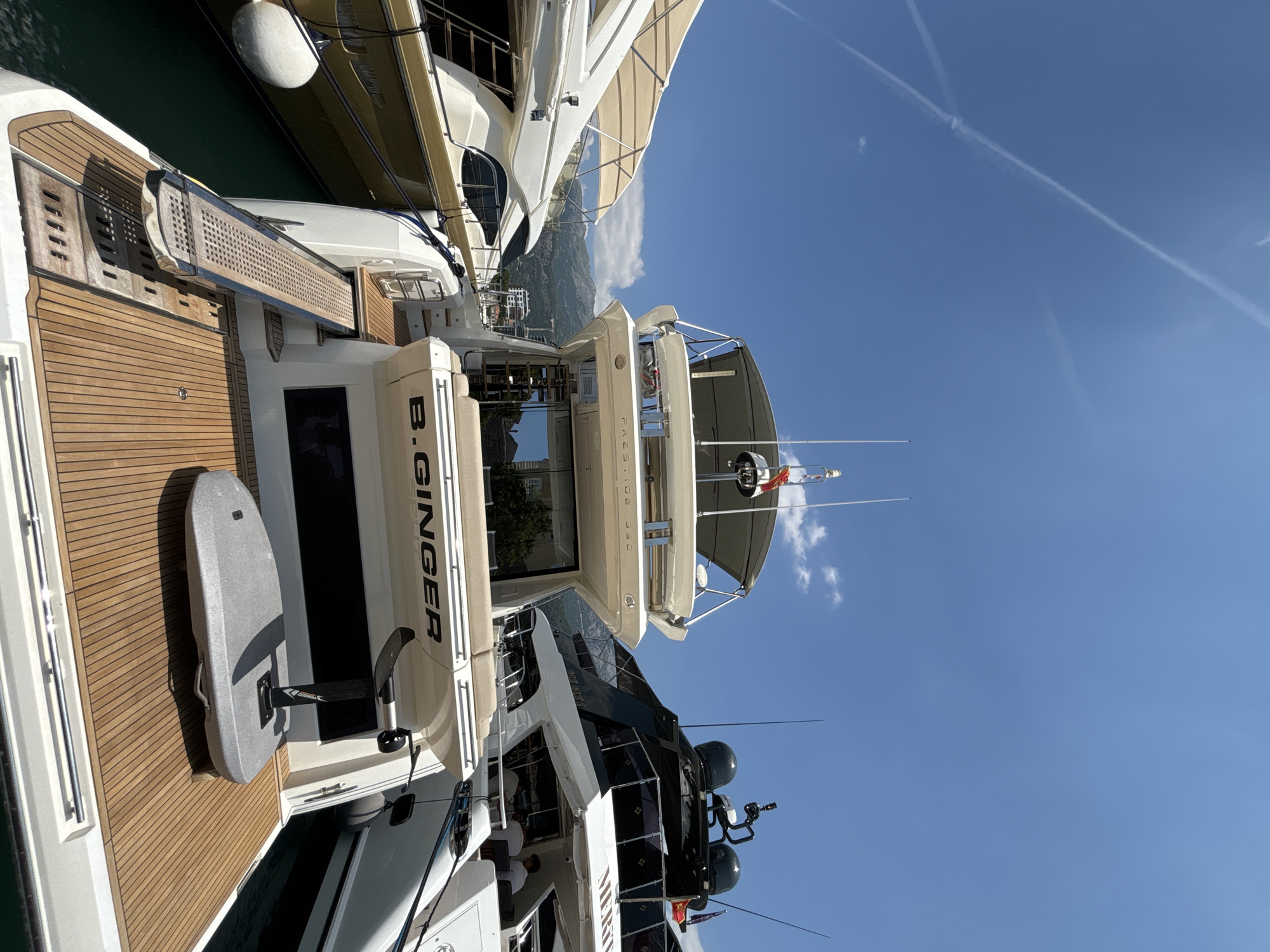 The image shows a rear view of the motor yacht "B. GINGER" docked in Budva, Montenegro, on a sunny day. The yacht has a white hull with teak decking at the stern. A retractable ramp extends from the stern to the dock. In the background, other yachts are docked, and mountains are visible in the distance under a clear blue sky with contrails. The overall scene depicts a luxurious marina environment.