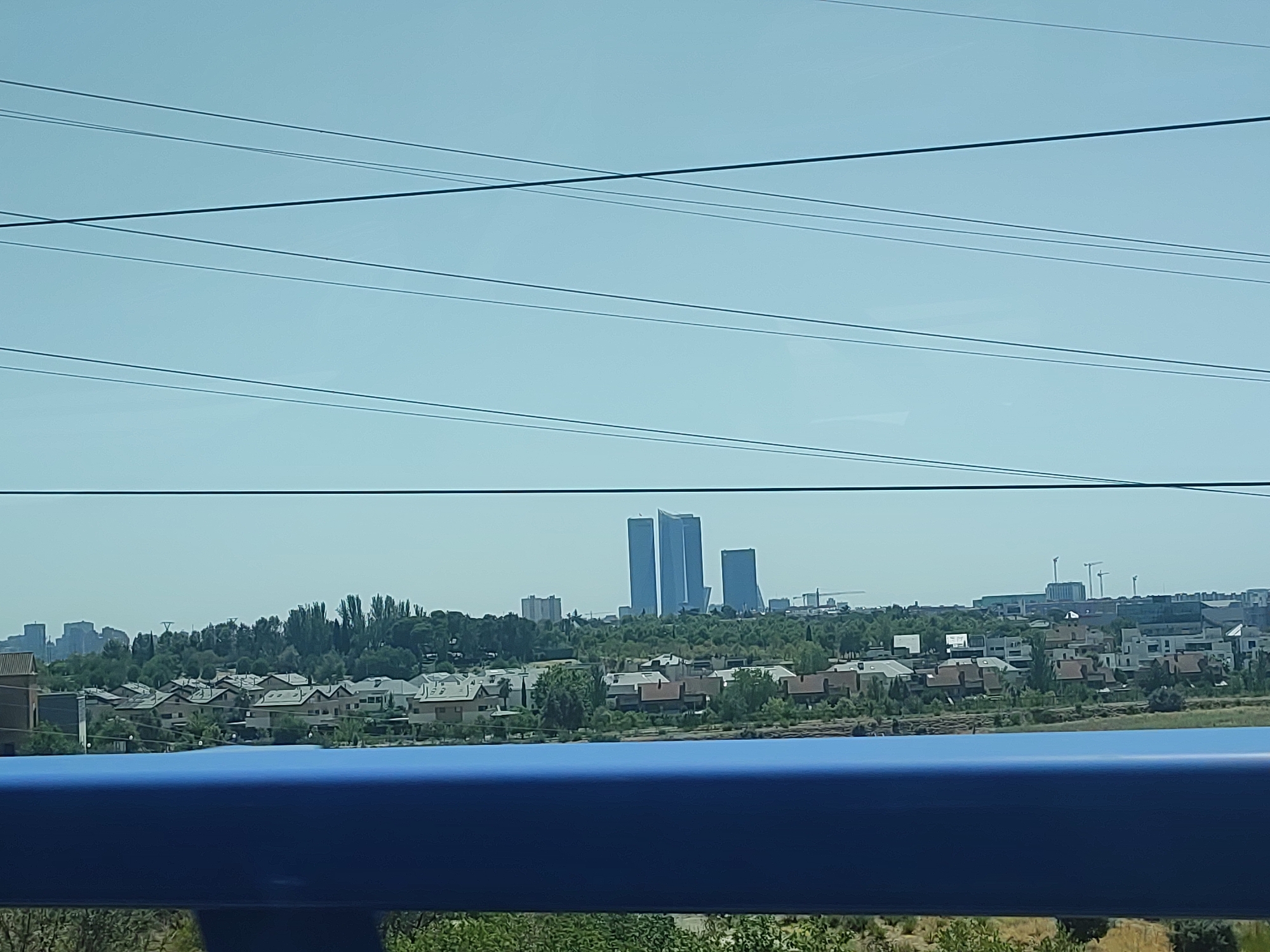 The image shows a panoramic view of a suburban area in Madrid, Spain, with a backdrop of the city skyline. In the foreground, a blue railing or barrier is visible, suggesting the viewpoint is from a bridge or elevated structure. Beyond the railing, a dense cluster of residential buildings with red-tiled roofs stretches across the mid-ground. These houses appear to be of a uniform design, indicative of a planned community or development.
Behind the residential area, a line of trees forms a natural boundary, with a variety of deciduous and evergreen species. Further in the distance, the distinctive skyscrapers of Madrid's financial district rise above the trees. The most prominent structures are three unusually shaped, tall buildings that appear to be part of the Cuatro Torres Business Area. To the left and right of these central towers, other, less distinct buildings and construction cranes are visible, hinting at ongoing development or the presence of other business complexes.
The sky is a clear, pale blue, suggesting a daytime, likely sunny, scene. Several power lines are strung across the upper portion of the image, intersecting the sky and the distant cityscape. The overall impression is one of a modern urban periphery, where residential zones meet commercial and business centers. No people are visible in the frame.