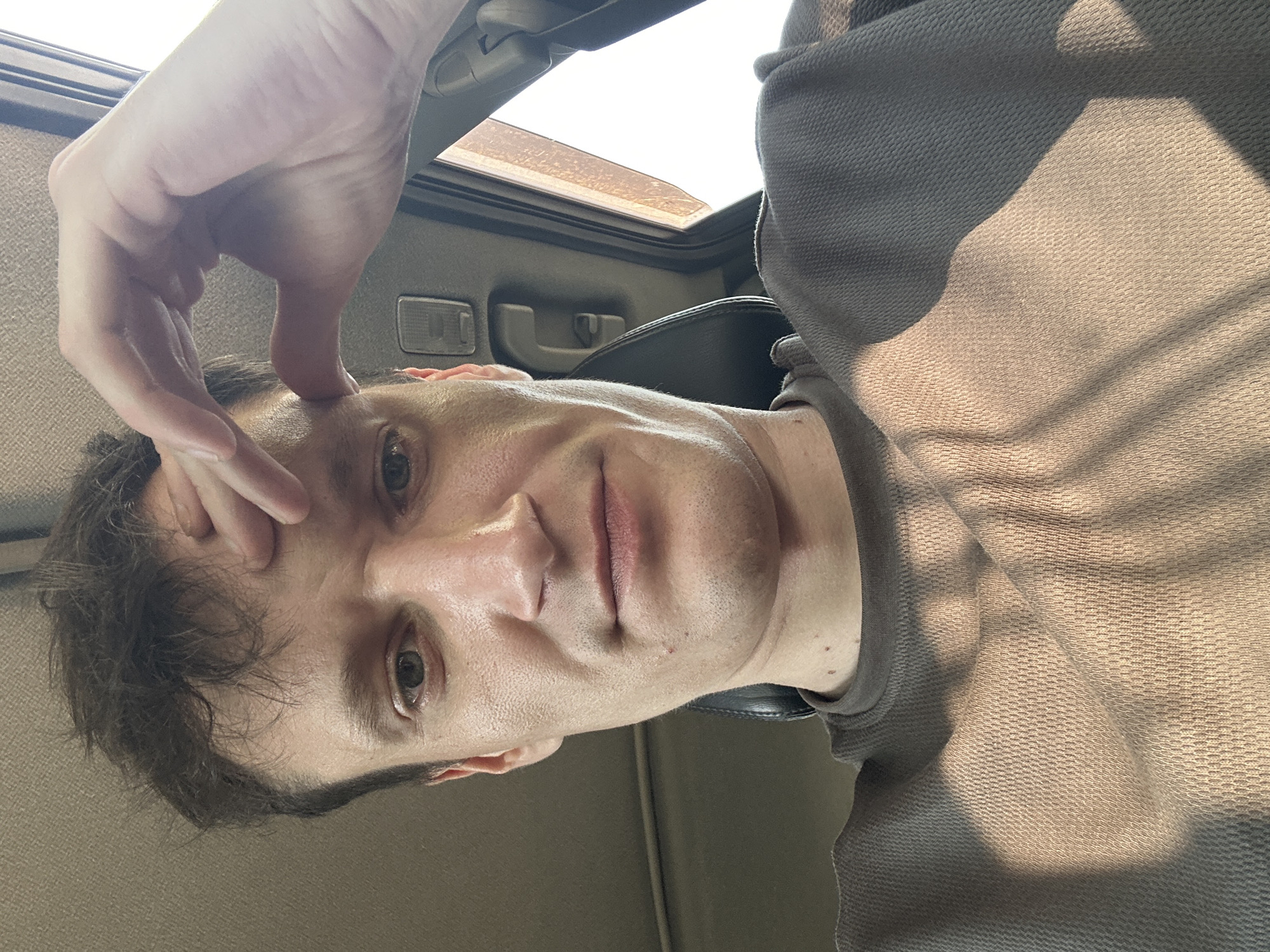A man is taking a selfie in a car. He is leaning his forehead on his hand, with his fingers splayed and touching his temple. He is wearing a brown crewneck shirt. The interior of the car is visible, including a headrest, a grab handle, and a sunroof. The lighting suggests it is daytime. The location is not specified in the image.