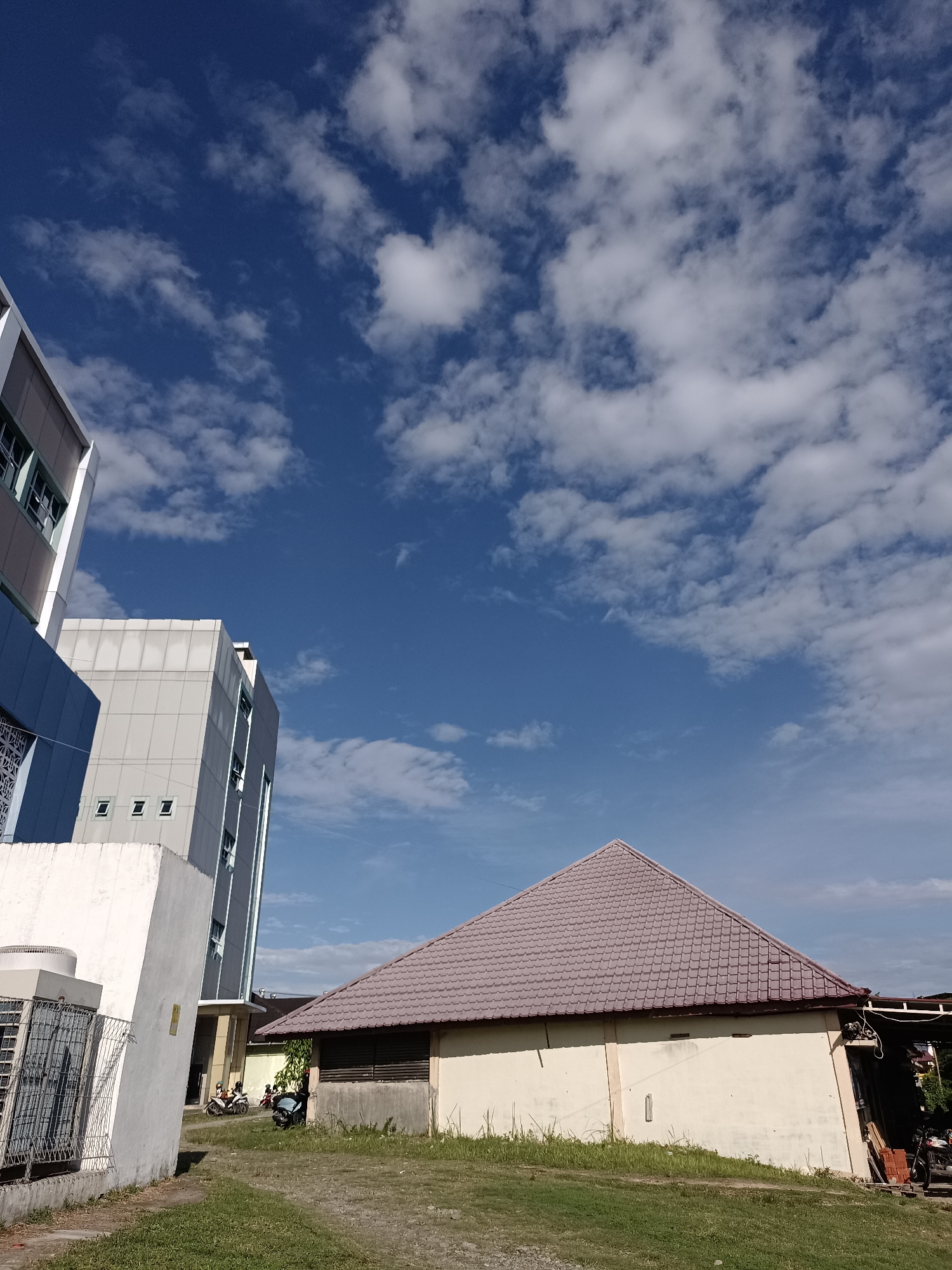The image depicts an outdoor scene under a bright blue sky with scattered white clouds. In the foreground, a light-colored, rectangular structure with a reddish-brown tiled pyramidal roof is visible. To the left, a portion of a tall, modern building with gray and blue exterior panels and visible windows rises into the frame. In front of the modern building, a white wall and a metal fence enclosure are present. To the right of the tiled-roof structure, part of another building or overhang is visible, with some wooden materials and a motorcycle parked. Several motorcycles and a few people are present in the middle ground, near the entrance to the tiled-roof structure. The ground is covered with grass and gravel. The scene appears to be a public area or an institutional setting within the city of Mesjid Reuntoh, Indonesia.