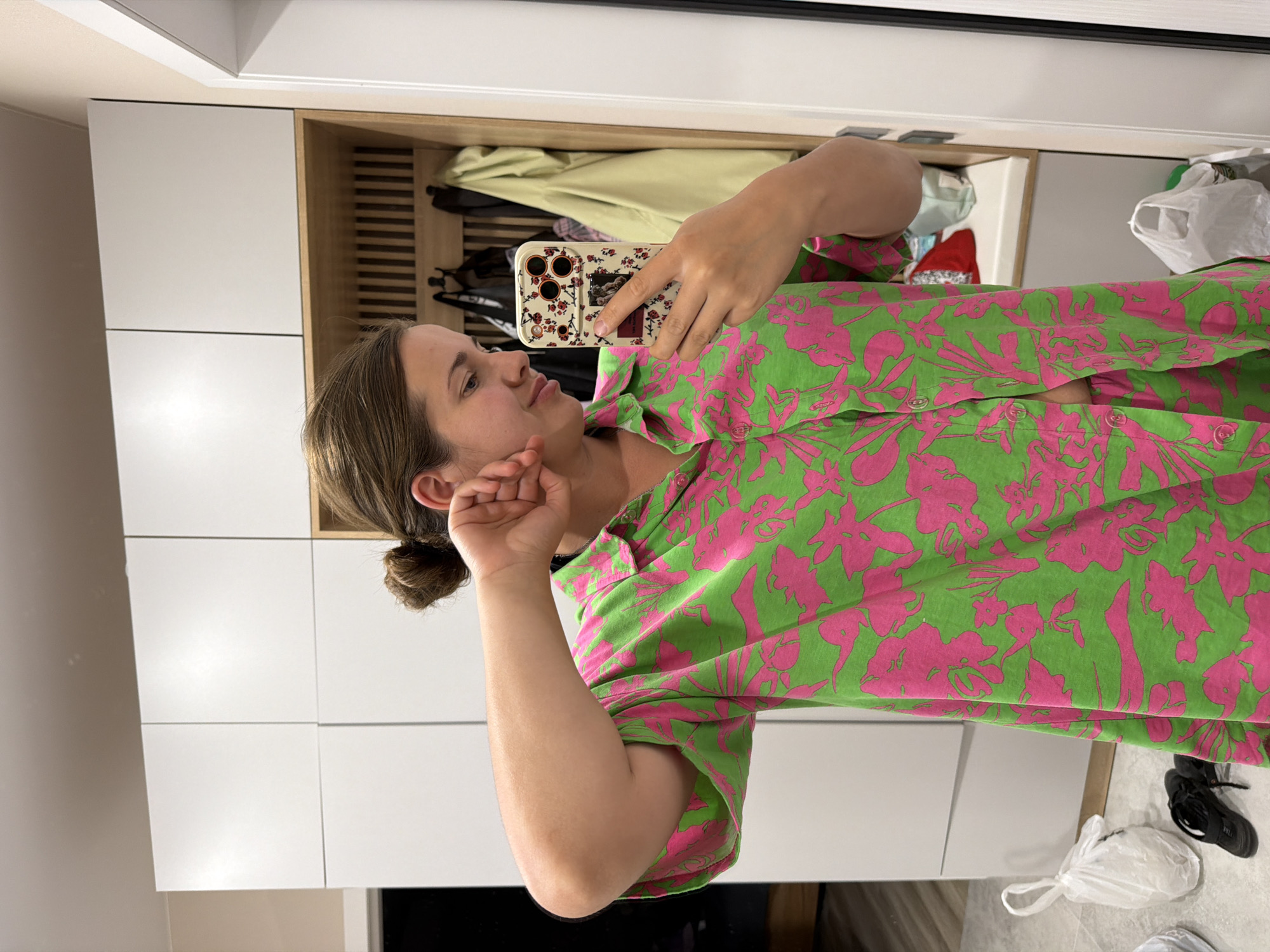 A woman is taking a selfie in her home, possibly in an entryway or closet area, in Novorossiysk, Russia. She has her hair in a bun and is wearing a green and pink floral print pajama set. She is holding a phone with a floral case in her right hand and her left hand is near her face. Behind her is a wall with white cabinets and a wooden shelving unit with clothes hanging on hooks. There are also plastic bags and shoes on the floor in the foreground.