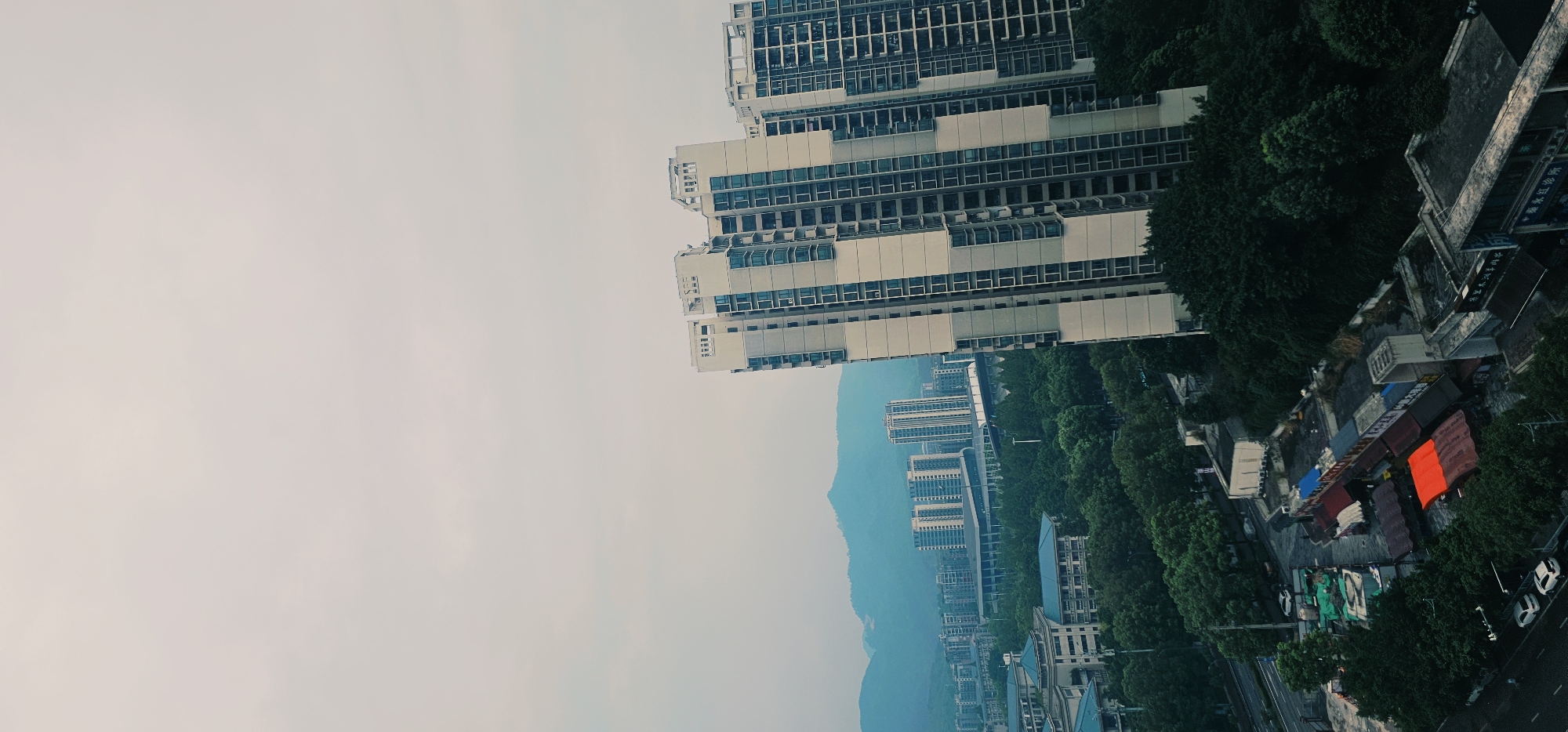 The image captures an aerial view of a densely populated urban area, likely in the Chongqing High-tech Zone, China. In the foreground, a large, modern residential building with a pale facade and numerous blue-tinted windows dominates the right side of the frame. The building appears to be multiple stories high, characteristic of high-rise apartments. To the left and in the mid-ground, a dense canopy of dark green trees forms a significant portion of the landscape, interspersed with more apartment buildings of varying heights and architectural styles, some featuring blue roofs. A prominent elevated highway structure, possibly a viaduct or monorail track, runs through the mid-ground, connecting different parts of the city. In the background, a range of misty, blue-hued mountains forms the horizon, suggesting a location with natural topography. Below the trees and closer to the viewer, there are some smaller, more utilitarian structures, including what appear to be temporary market stalls or workshops with corrugated metal roofs, one of which is brightly colored in red. Several white cars are visible on a road in the lower left portion of the image. The sky is overcast and a pale gray, indicating a cloudy day. There are no people clearly visible in the image.