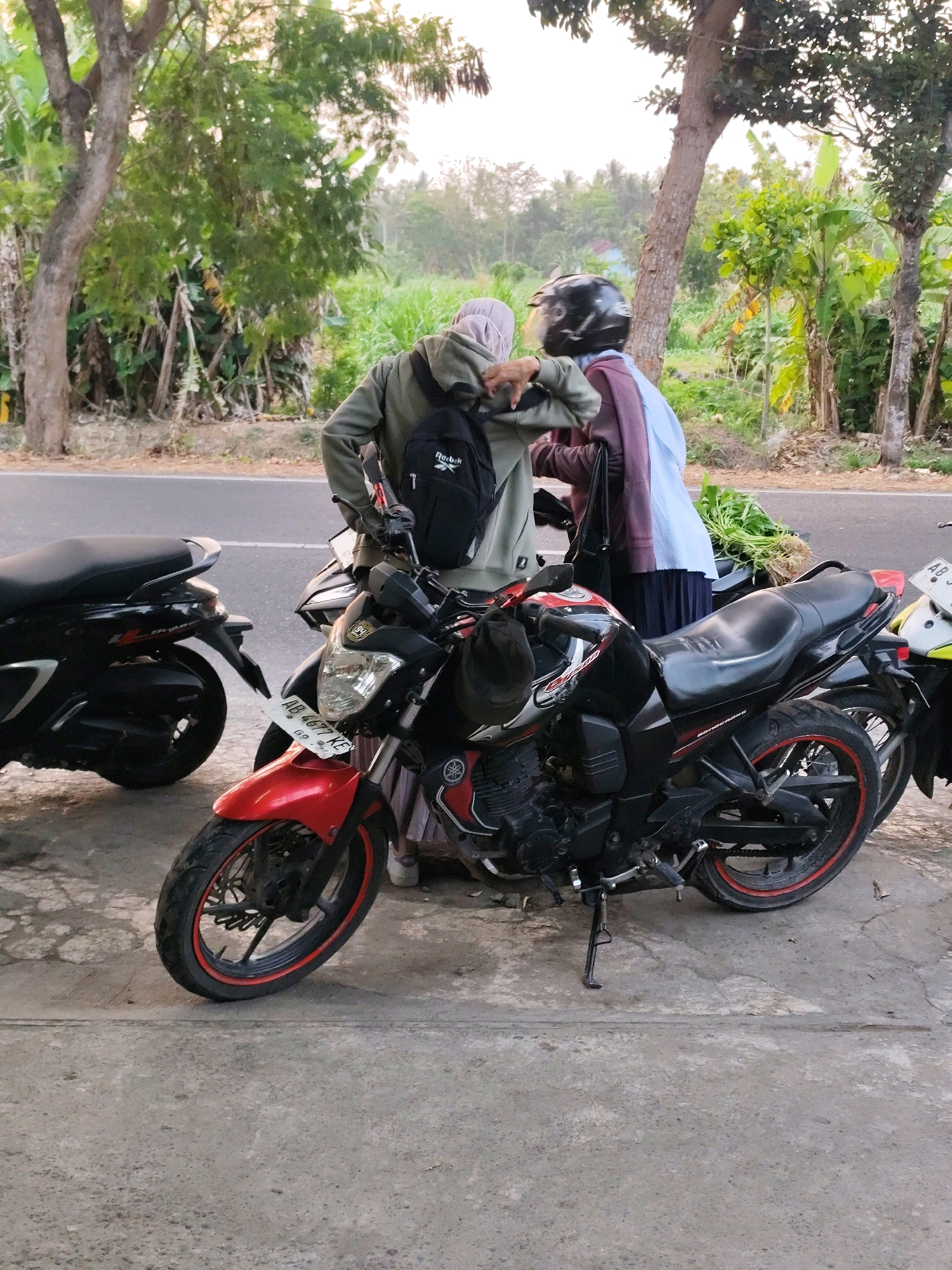 The image depicts two individuals standing next to parked motorcycles on a concrete surface adjacent to a road. The environment suggests a rural or semi-rural setting with trees, vegetation, and distant structures visible.
One individual, wearing a light green hooded jacket and a black backpack, is facing away from the viewer, leaning towards a black motorcycle. The backpack has a "Reebok" logo. The second individual, wearing a dark maroon jacket over a light blue garment and a dark skirt, is partially obscured, also interacting with the same motorcycle. A black helmet is placed on the seat of the motorcycle. A bunch of green vegetables, possibly harvested produce, is placed on the back of the black motorcycle.
In the foreground, a red and black motorcycle is parked. Its front wheel is visible, featuring a black rim with a red accent stripe. A license plate with the inscription "AB 4677 KE" is attached to the front fender. To the left, a black scooter is parked. The ground is a weathered concrete surface with visible cracks and patches. The background shows a road, trees, and foliage. The overall lighting suggests it might be late afternoon or early morning. The location is specified as Bambanglipuro, Indonesia.