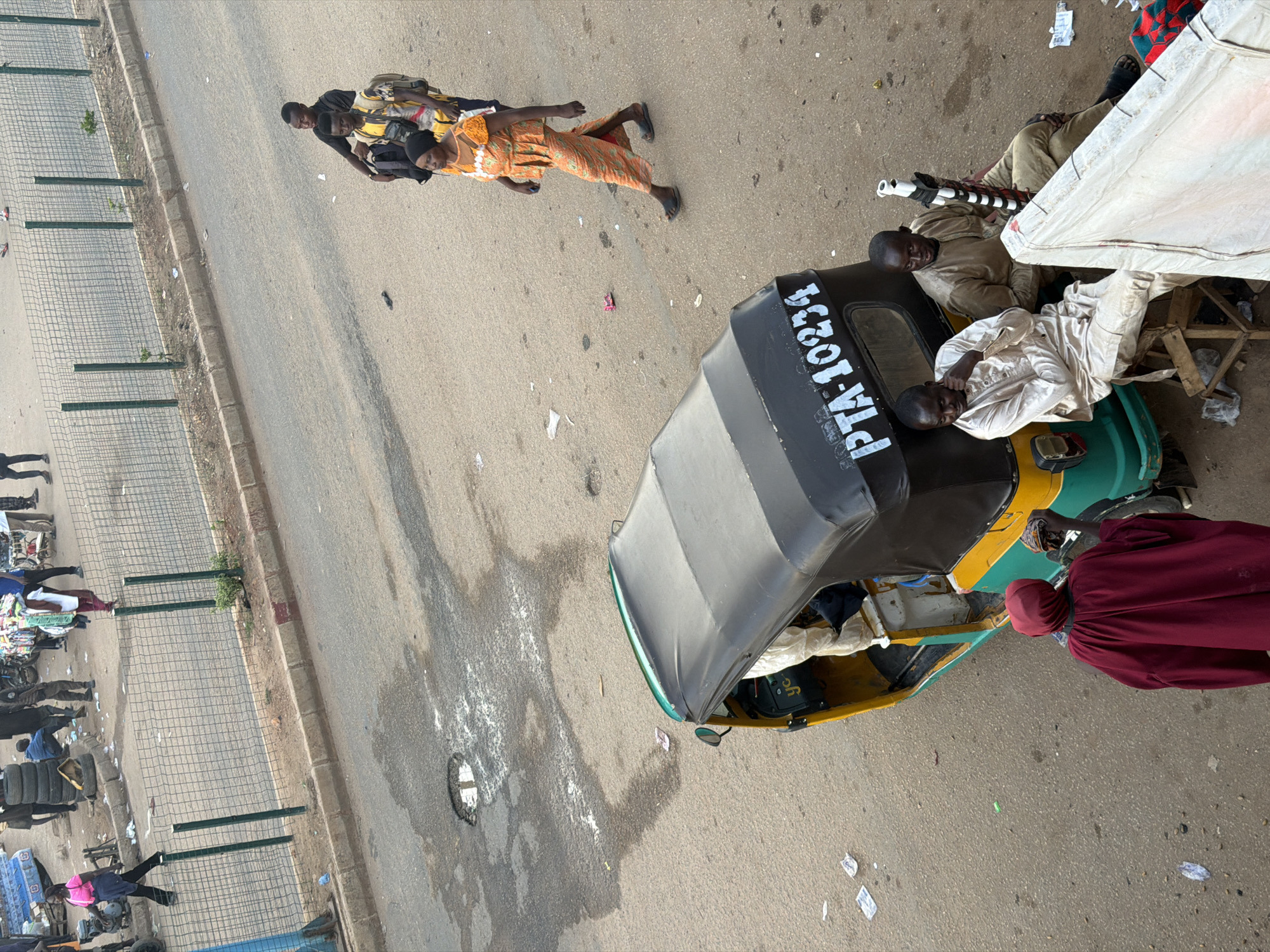 An overhead, medium shot captures a busy street scene in Bukuru, Nigeria. In the foreground, a green and yellow auto-rickshaw, identified by the license plate "PTA-10234," is parked on the asphalt road. Three men are near the auto-rickshaw: one in a beige robe sits on a wooden stool next to the vehicle, while two others, one in a beige outfit and another in a tan outfit, are seated on a white tarp to the right. The man on the left is leaning on his hand, looking down, while the man on the right is holding a white and black striped object and looking towards the left.
Further back, several people are walking on the street and sidewalk. A woman in a bright orange patterned dress and black sandals walks towards the right, carrying a black bag on her left shoulder. Behind her, two men with backpacks walk away from the camera, one in a yellow and black shirt and the other in a black shirt. On the far left, a woman in a pink t-shirt and black pants walks away from the auto-rickshaw.
The environment appears to be a market area, with various objects and debris scattered on the road and sidewalk. A metal fence runs along the side of the road, separating it from a dirt and gravel area. In the background, more people and vehicles are visible, suggesting a bustling urban environment. The road itself has several potholes and patches of what appears to be dried white liquid.