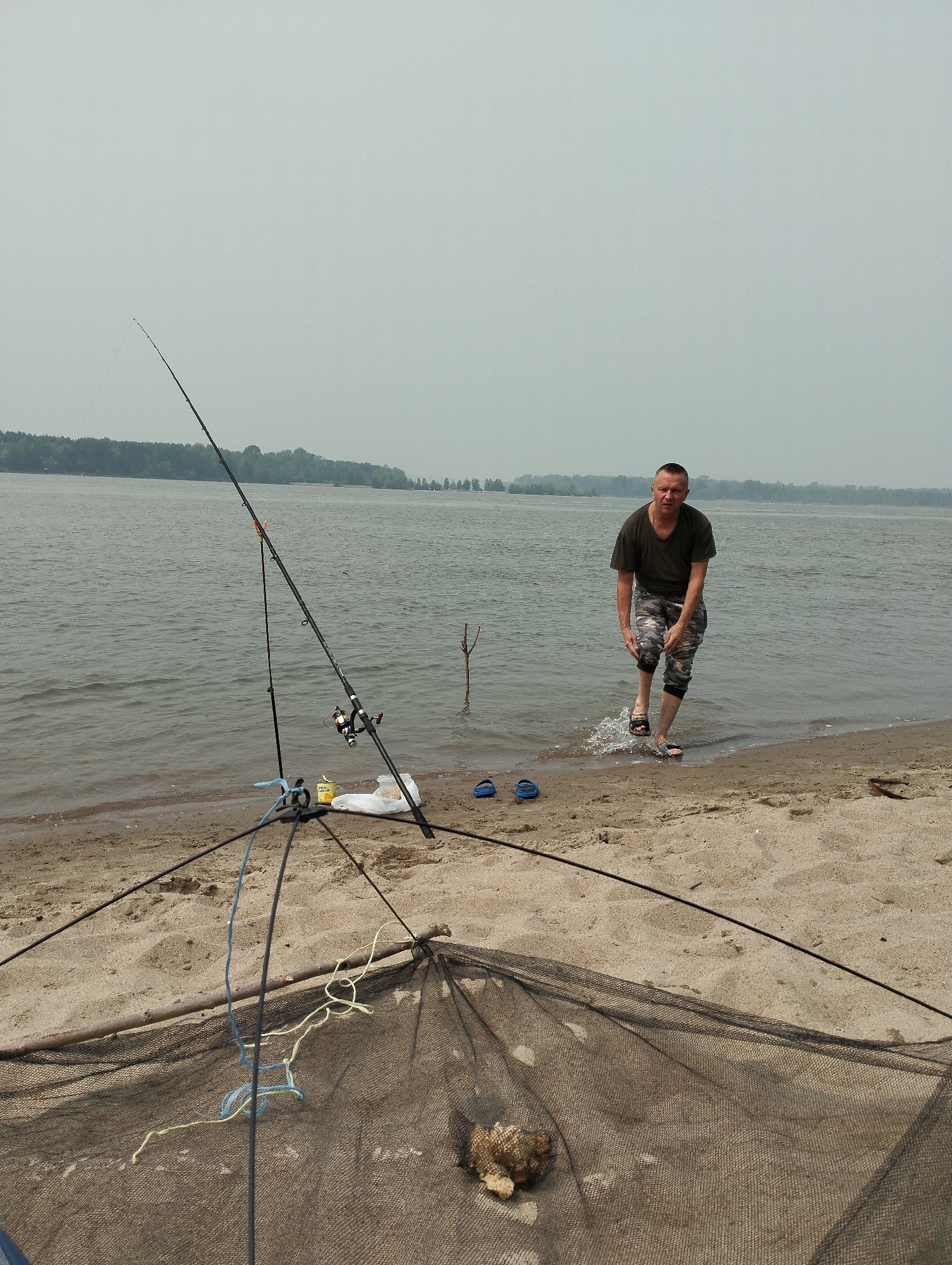 A man stands in shallow water on a sandy riverbank in Bibikha, Russia, with fishing equipment set up on the shore. The man, wearing a dark green t-shirt and camouflage-patterned shorts and sandals, is mid-stride, his legs creating splashes as he moves from the water onto the sand. To his right, two blue flip-flops lie on the beach. In the foreground, two fishing rods are propped up by a tripod stand. One rod is taller and thinner, with a fishing line extending upwards. The second rod is shorter and thicker, with a fishing reel attached. A fishing net lies spread out on the sand in front of the tripod, with a piece of driftwood and some cordage visible near its center. A small yellow tin and a white plastic bag are positioned behind the tripod. The background consists of a wide, calm river and a distant, tree-lined shore under an overcast sky. The overall scene suggests a fishing excursion.