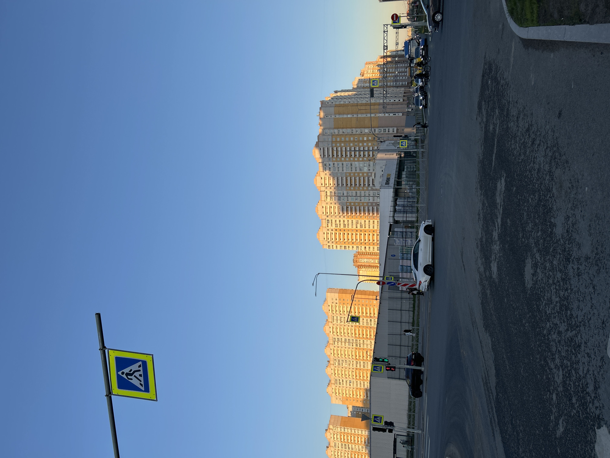 The image depicts a cityscape in Saint Petersburg, Russia, featuring a pedestrian crossing sign in the foreground set against a clear blue sky. Adjacent to the sign is a row of high-rise apartment buildings, reflecting the warm light of either sunrise or sunset. A busy street runs alongside the buildings, populated with cars, traffic lights, and pedestrian signs. The scene suggests an urban environment with typical infrastructure and modern residential architecture.