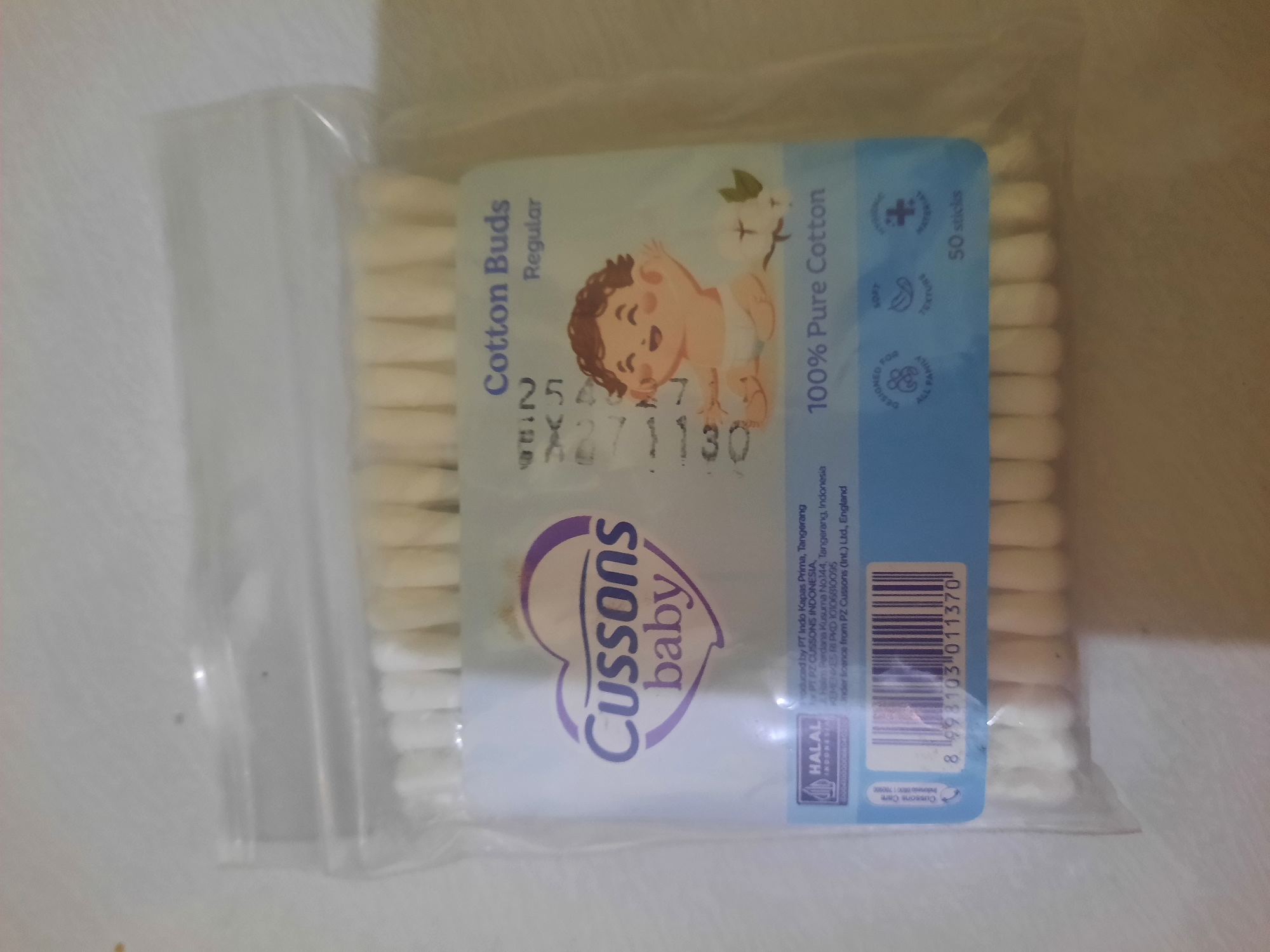 The image shows a sealed plastic package of Cussons baby cotton buds. The package is mostly transparent, revealing rows of white cotton buds inside. The front of the package features the Cussons baby logo, which is a stylized purple heart with the text "Cussons baby" inside. Below the logo, it states "Cotton Buds Regular" and "100% Pure Cotton". There are also small icons indicating the product is "Designed for All Family", has a "Soft Texture", and is made of "Hygienic Material". The package also includes a barcode and the text "50 sticks". A HALAL Indonesia logo is visible in the lower left corner. The image is a top-down, slightly angled shot of the product. The lighting is somewhat dim, with some reflections on the plastic packaging. The background is a plain, light-colored surface, possibly a table or countertop. There is no indication of the city of Lhokseumawe, Indonesia, in the image.