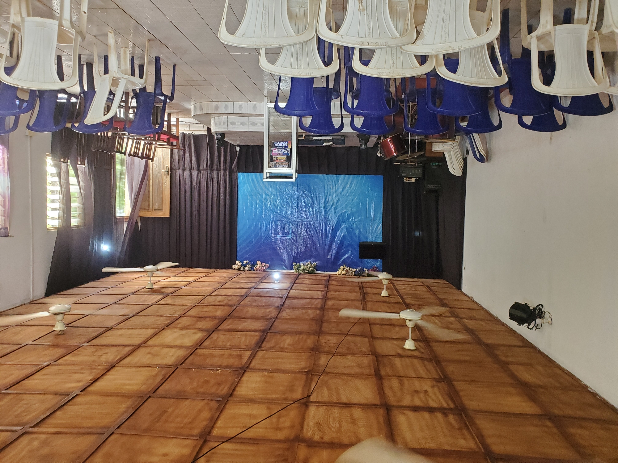 The image depicts the interior of what appears to be a church or community hall in Oturkpo, Nigeria. The room is furnished with rows of blue and white plastic chairs, facing a stage area. The stage features a large blue tarp backdrop, a keyboard, a drum set, and a podium with literature. The ceiling is paneled with wood and equipped with ceiling fans. Stage lighting equipment is visible on the walls and stage. The overall impression is of a functional, somewhat sparsely decorated space prepared for an event or service.