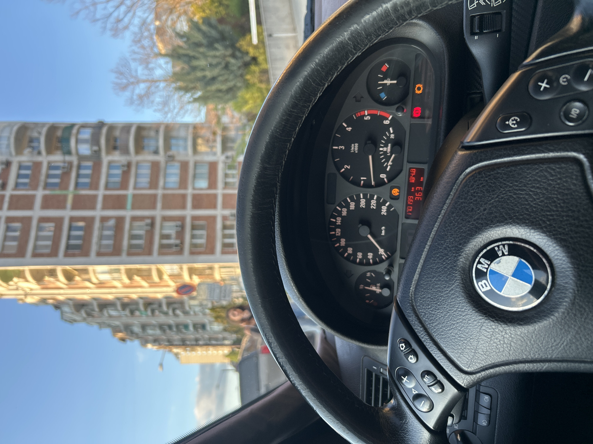 The image depicts the interior of a BMW, focusing on the steering wheel and dashboard. The BMW logo is prominently visible on the steering wheel. The dashboard displays the speedometer, tachometer, and other gauges, with various warning lights illuminated. Through the windshield, an out-of-focus view of a building and street scene can be seen. Based on the architectural style of the building and the street signs visible, it is likely the location is in the city of Sofia, Bulgaria. The sky is blue and partially visible through the windshield. There is also an out of focus person visible in the reflection on the windshield.