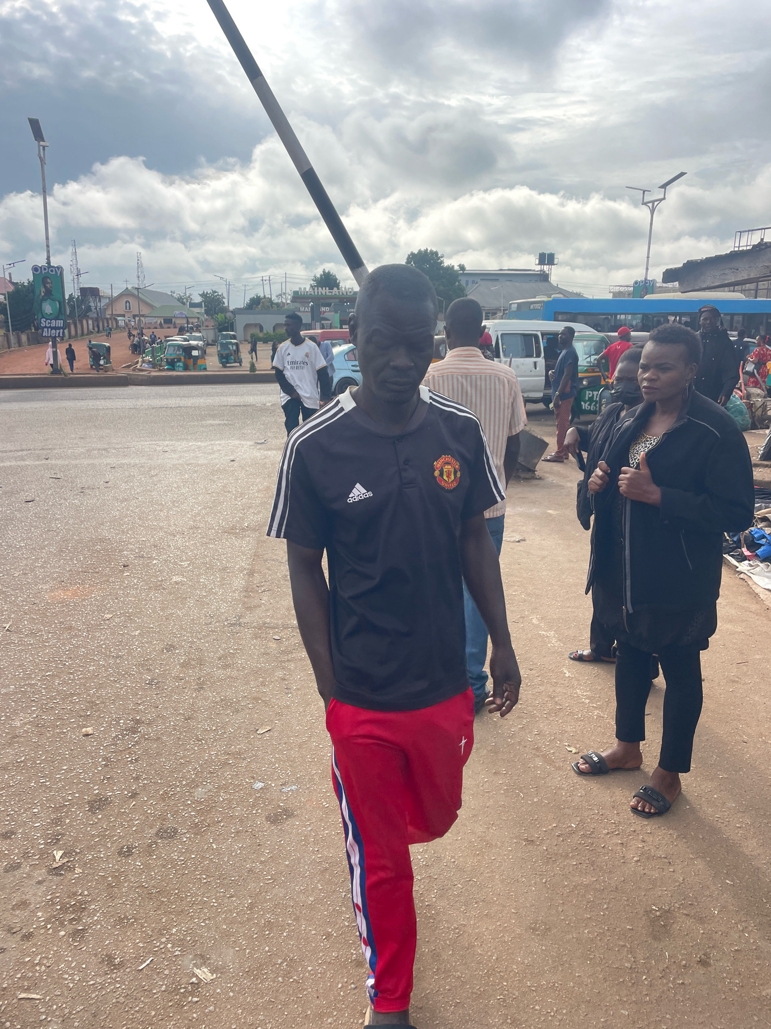 The image depicts a street scene in Gura Top, Nigeria, under an overcast sky. In the foreground, a man wearing a black Adidas t-shirt with white stripes on the sleeves and red track pants with white and blue stripes walks towards the camera. He has his left hand in his pocket. Behind him, a woman in a black jacket and patterned top stands, holding her jacket. Further back, other individuals and various vehicles, including tricycles and a blue bus, are visible. A billboard with a "Scam Alert" message is also present. The ground is a dusty, granular surface, characteristic of many Nigerian roads. The overall impression is that of a busy urban intersection or market area.