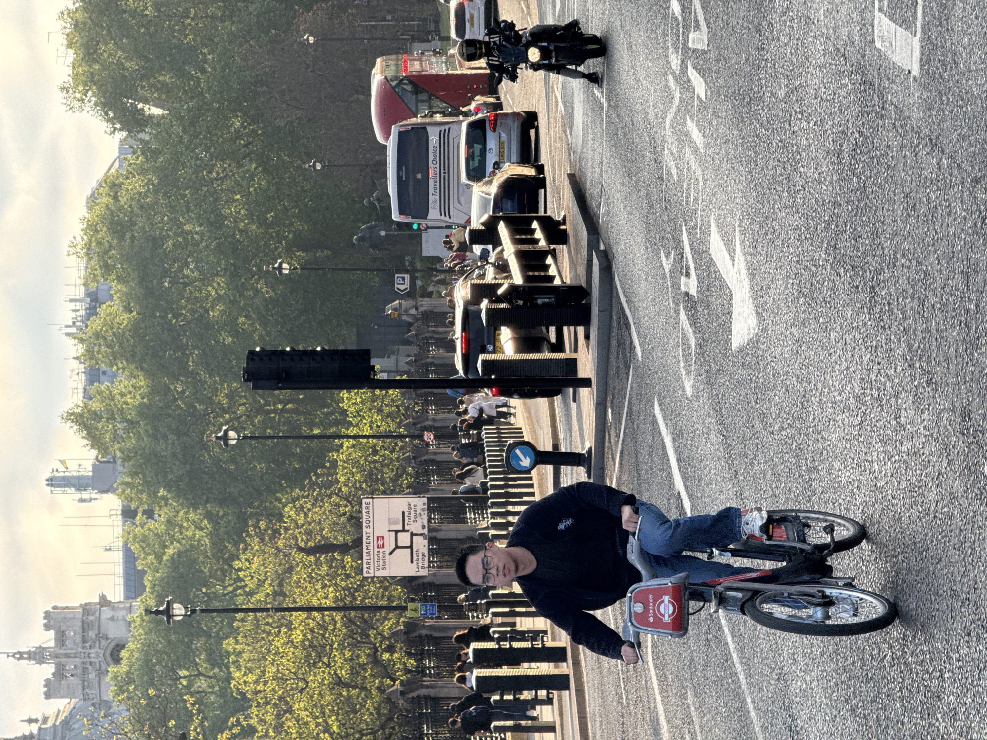 The image shows a person riding a Santander Cycles rental bike on a street in London, United Kingdom. In the background, there is a bus, cars, and a motorcycle, indicating traffic on the road. Street signs indicate that the location is near Parliament Square, Victoria Station, Lambeth Bridge, and Trafalgar Square. There are pedestrians walking on the sidewalk alongside trees and street lamps. A building is seen beyond the trees. The rider faces the camera.
