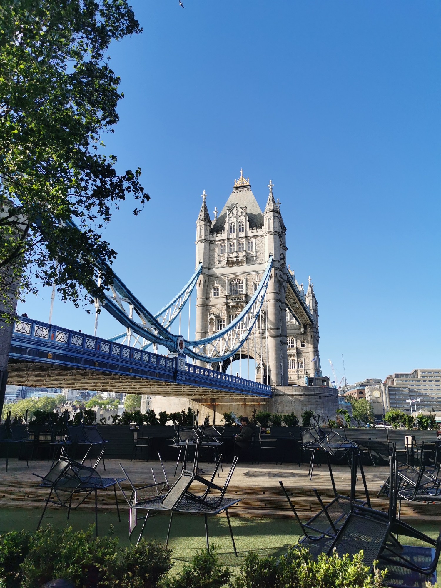 This vibrant daytime image captures an iconic London landmark, Tower Bridge, framed by lush greenery and an outdoor seating area. The scene unfolds on a clear, sunny day, indicated by the brilliant blue sky overhead.
In the foreground, an outdoor terrace, likely belonging to a cafe or restaurant, is visible. It features a wooden deck and patches of artificial turf. Numerous black metal chairs are scattered across this area, many of them stacked or overturned, suggesting the venue might be closed or undergoing setup/cleanup. One person is seated at a table on this terrace in the mid-ground, appearing to be relaxing or smoking.
Dominating the mid-ground is the majestic Tower Bridge. Its intricate stone towers, with their distinctive Gothic architecture, rise prominently against the sky. The bridge's famous blue and white suspension cables and girders are clearly visible, with some gold-colored crests adorning the blue framework. Small figures of people can be seen walking on the bridge's upper walkway. A large, leafy green tree partially frames the bridge on the left side of the image.
In the background, distant city buildings and construction cranes are visible, hinting at the bustling metropolis. Beneath the arch of the bridge on the right, the outline of the historic Tower of London can be glimpsed, adding another layer of historical context to the scene. A small bird is also visible soaring in the clear blue sky in the upper left quadrant. The overall atmosphere is serene and bright, capturing a quiet moment beside one of London's most recognizable structures. There is no clearly legible text within the image.