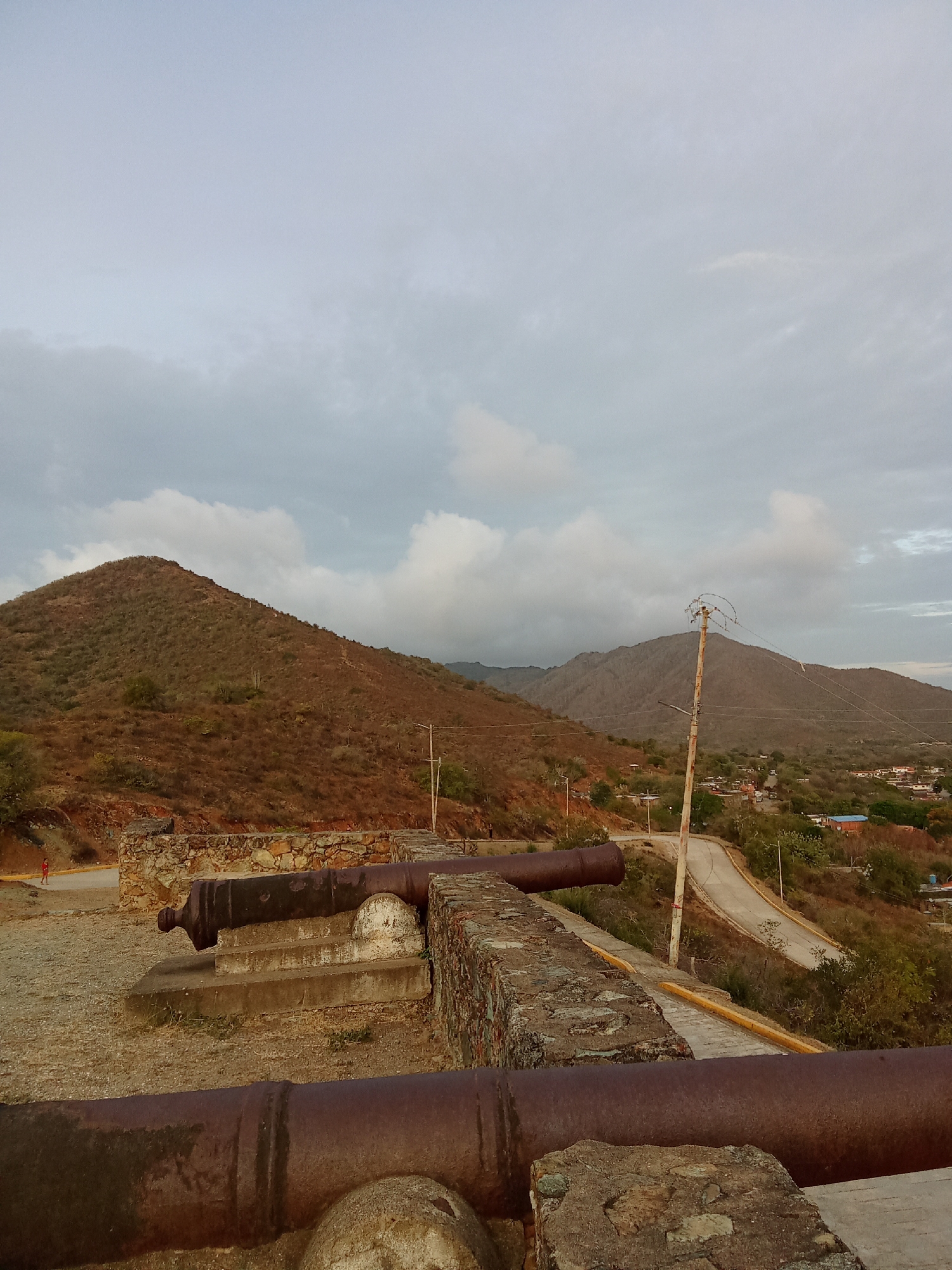 The image depicts a historical site in Santa Ana, Venezuela, featuring cannons overlooking a landscape. Two large, rusted cannons are in the foreground, mounted behind a low stone wall. In the background, a road winds through a hilly landscape covered in dry vegetation. A telephone pole stands along the roadside. The sky is overcast with gray and white clouds. To the left, a small red figure can be seen, likely a person walking on the road. Further in the background, there are buildings, suggesting a small town or residential area nestled among the hills.