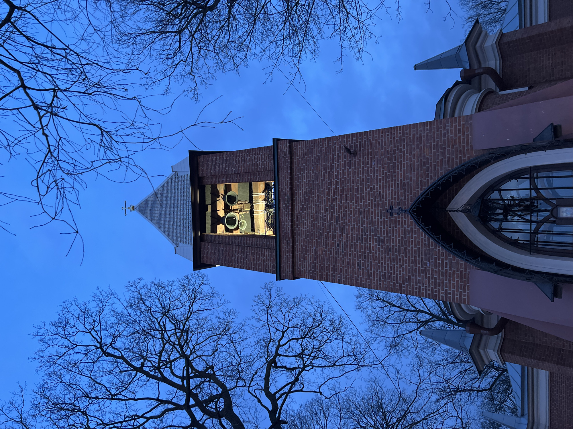 A low-angle, exterior photograph captured during twilight depicts the bell tower and upper facade of the Evangelical Lutheran Church of Saint Michael, located at Sredny Prospekt 18, Vasilyevsky Island, Saint Petersburg, Russia. The central feature is a tall, reddish-brown brick bell tower, topped with a gray, conical roof that culminates in a white cross. Below the roof, the bell chamber is illuminated from within, showcasing two large, dark-colored bells and one smaller bell. The tower's lower section transitions into a pink-hued base, featuring a large, dark-framed arched window or entrance with a decorative grille. Adjacent structures flank the main tower, exhibiting similar brickwork and topped with smaller, pointed gray spires. The background consists of a deep blue sky, against which the intricate, bare branches of deciduous trees are silhouetted on both sides of the church. A thin, dark cable is visible traversing the sky horizontally in the mid-ground. The scene is devoid of people, actions, or interactions.