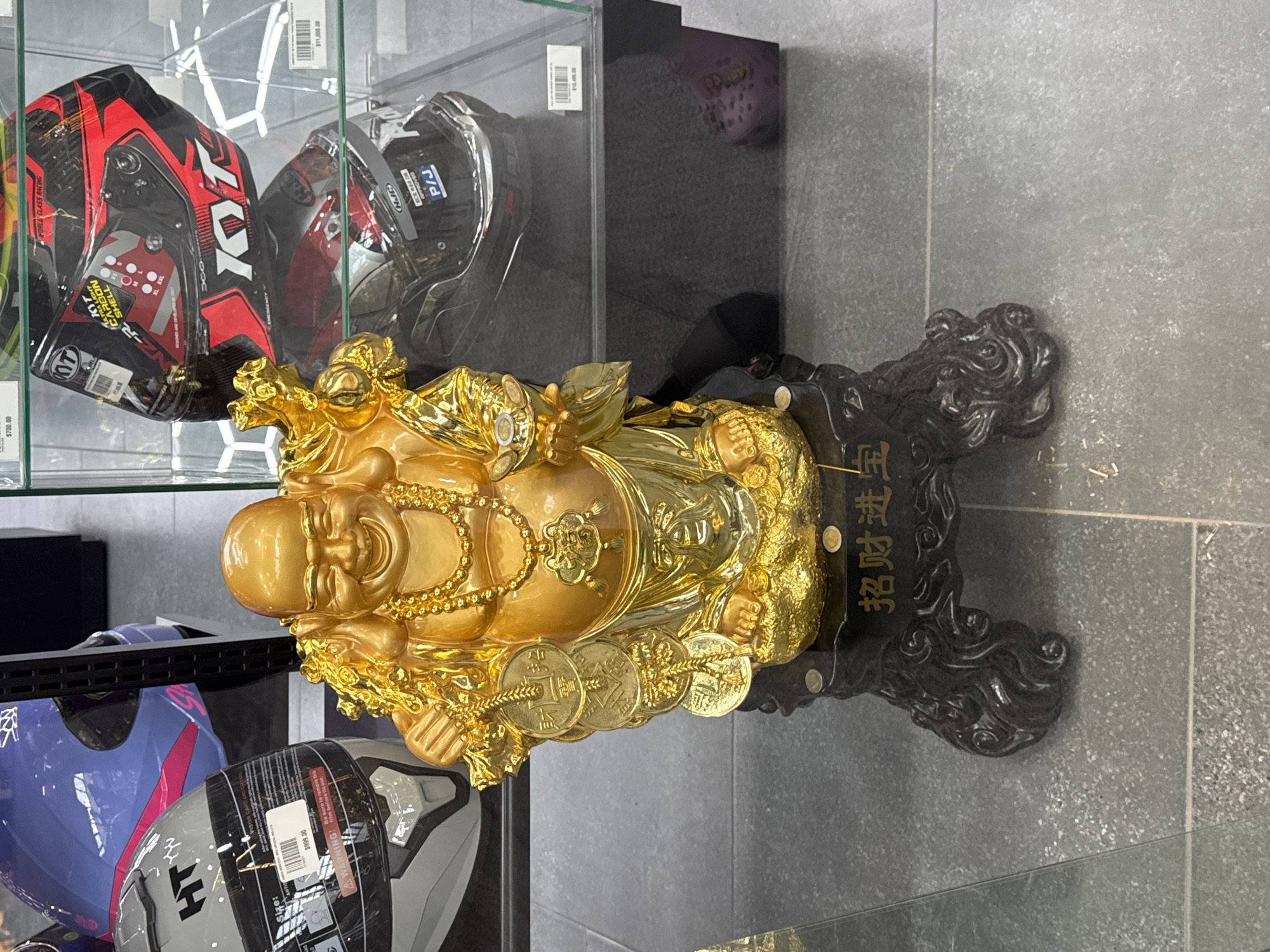 Here is a detailed description of the image:
**General Description:** The image shows a golden Buddha statue on a black ornate stand, positioned in front of a display case containing motorcycle helmets.
**Objects:**
* **Golden Buddha Statue:** A prominent golden-colored statue of Buddha with a smiling face. It is adorned with beads and is holding what appears to be coins.
* **Black Ornate Stand:** The statue is placed on an intricately designed black stand. Chinese characters are visible on the stand.
* **Motorcycle Helmets:** Several motorcycle helmets are visible in the display case behind the statue. They are of various colors, including red, black, blue and grey. Some helmets have visible brand logos.
* **Display Case:** The helmets are kept inside a glass display case.
**Environment:**
* **Indoor Setting:** The setting is indoors.
* **Flooring:** Gray tiled flooring is visible.
**Overall Impression:** The image conveys a commercial setting, possibly a store that sells motorcycle helmets and other related accessories. The presence of the golden Buddha suggests a cultural or decorative aspect within the store.
Based on the tags $700.00 to $11,000.00 it is likely to be in Mexico. The tags on the helmets are in US Dollar Currency. The language on the base is likely to be Chinese, and the subject matter is Buddha. There is a large Chinese population in Puebla, Mexico and therefore this image is very likely to be set in Puebla, Mexico.