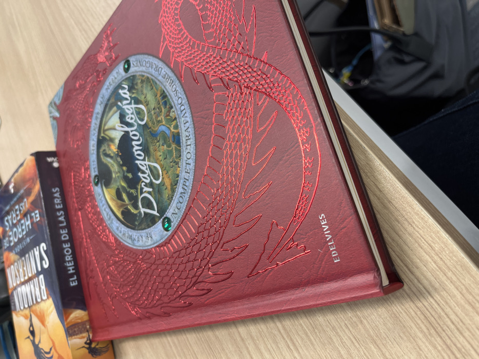 The image shows several books. A red hardback book titled "Dragonologia" features a detailed dragon illustration and the tagline "UN COMPLETO TRATADO SOBRE DRAGONES". The publisher "EDELVIVES" is also visible on its spine. Beside it is "EL HEROE DE LAS ERAS" by Brandon Sanderson, suggesting a fantasy genre. The books are placed on a light wooden surface next to a white object with dark fabric, possibly clothing.