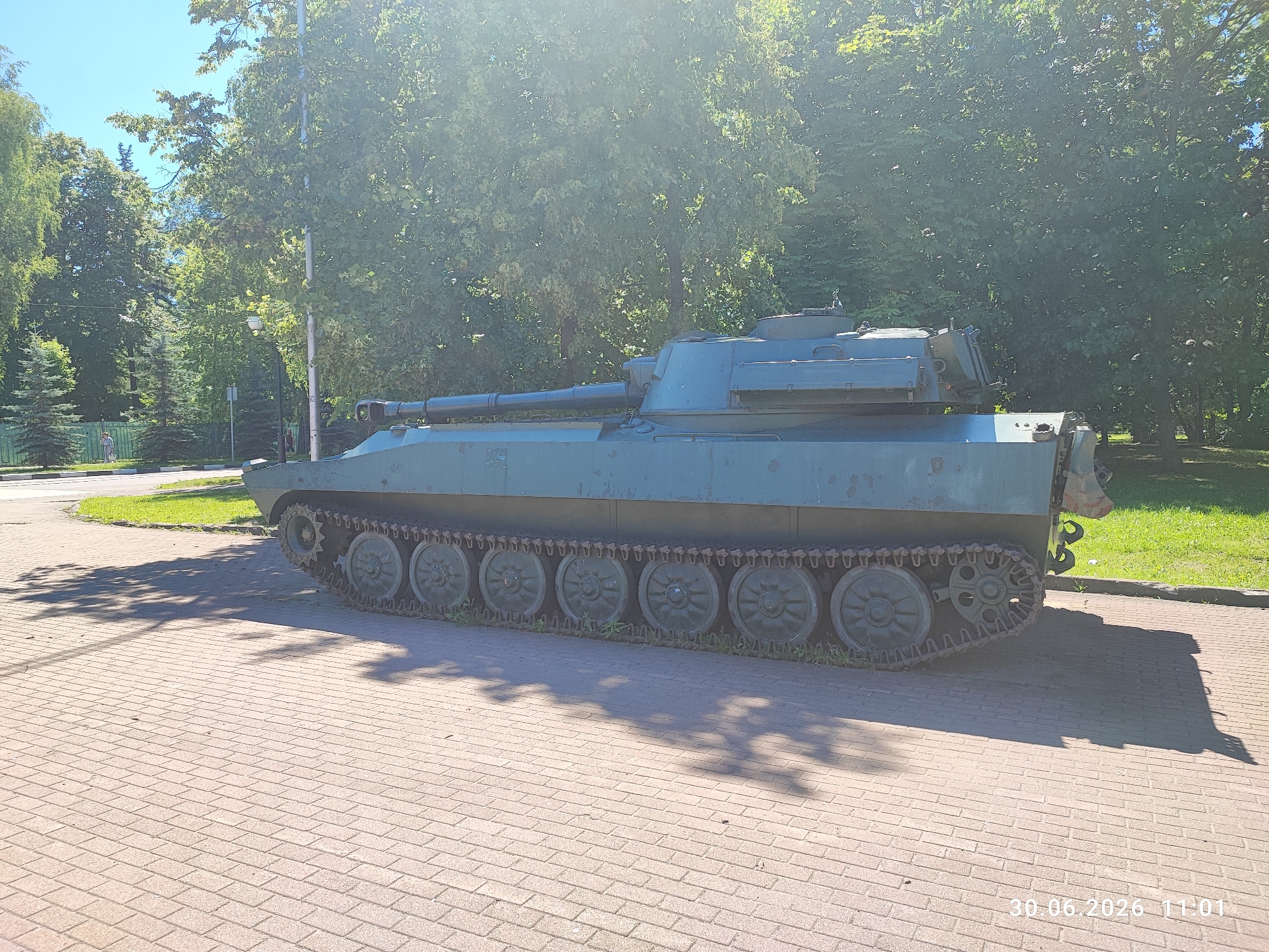This is a medium-shot, eye-level, exterior, horizontal photograph of a green armored vehicle, possibly a self-propelled gun or tank, parked on a paved area in what appears to be a park or public space. The vehicle is the primary subject, viewed from its left side, with its tracks, hull, and turret clearly visible. The gun barrel extends forward.
The setting is outdoors, with lush green trees and foliage in the background, suggesting a daytime scene on a sunny day due to the strong sunlight and shadows cast by the trees on the pavement. The sky is bright blue. The pavement is made of light-colored interlocking bricks. To the left, a glimpse of a road and a grassy area with a green fence can be seen. The overall atmosphere is peaceful and still, with no people visible in the frame.
There is text visible in the bottom right corner of the image: "30.06.2026 11:01". This indicates the date and time the photograph was taken, suggesting the image is a recent digital capture. The location context provided is Lytkarino, Russia.