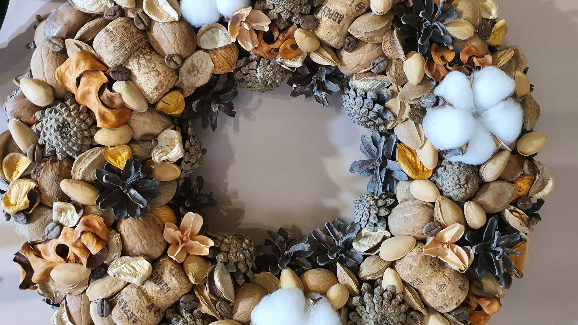 The image displays a circular wreath composed of natural elements such as nuts, pinecones, dried flowers, coffee beans, and cotton balls. The wreath is arranged on a neutral-colored, flat surface. There are no people or animals visible in the image, and no specific location within Krasnoyarsk, Russia is identifiable. The focus is solely on the decorative wreath.