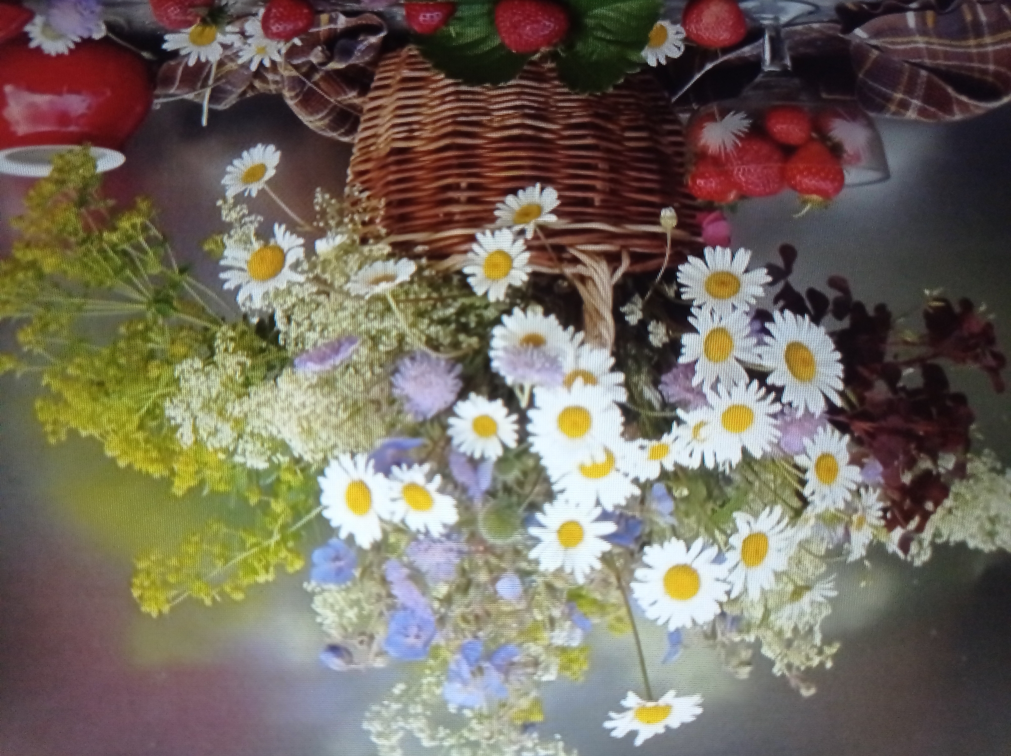 This is a still life image featuring a woven basket filled with a bouquet of wildflowers, primarily daisies and some purple flowers, with a sprig of yellow greenery. To the left of the basket, a glass compote dish holds several ripe strawberries. A red ceramic pot is visible to the right. The composition is set against a blurred, muted background. There is no discernible information about the city of Mukachevo, Ukraine, or any specific location within it, as the image depicts a still life arrangement. The scene is static and does not contain any people or animals.