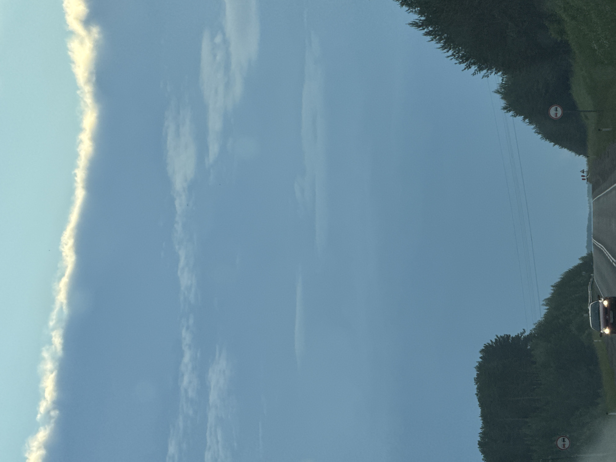 The image captures a road in the city of Karagay, Russia, under a vast, pale blue sky. A thin, luminous band of clouds stretches horizontally across the upper portion of the frame, catching the light. Below this, the sky is a uniform blue, with faint, wispy cloud formations scattered at various altitudes.
The road, paved and appearing to be a two-lane highway, curves gently into the distance. On either side of the road, dense green trees form a natural canopy, creating a sense of enclosure. The trees on the left are closer to the road and form a thick forest. On the right, the trees are also dense, with their branches reaching towards the road.
A red car is visible on the road, moving towards the viewer and emitting light from its headlights. Further down the road, in the distance, a group of small figures, presumably people, can be seen. There are two traffic signs visible: one on the left side of the road, indicating no overtaking, and another on the right side with the same prohibitory symbol. The overall scene suggests a journey through a natural, somewhat remote landscape during daylight hours, possibly late afternoon or early evening given the soft lighting.