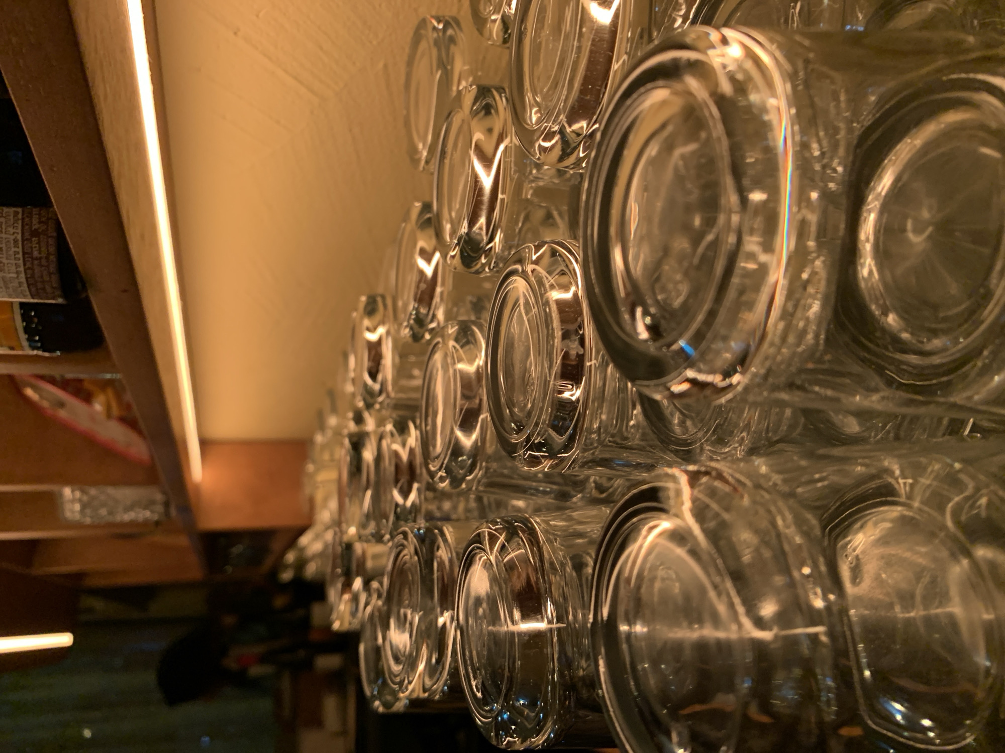A close-up, low-angle shot displays rows of clear glass cups stacked upside down. A warm, yellowish light illuminates the scene, highlighting the circular bases of the cups and creating reflections. To the left, a shelf with a blurred bottle is visible. The location is Tel-Aviv, Israel.