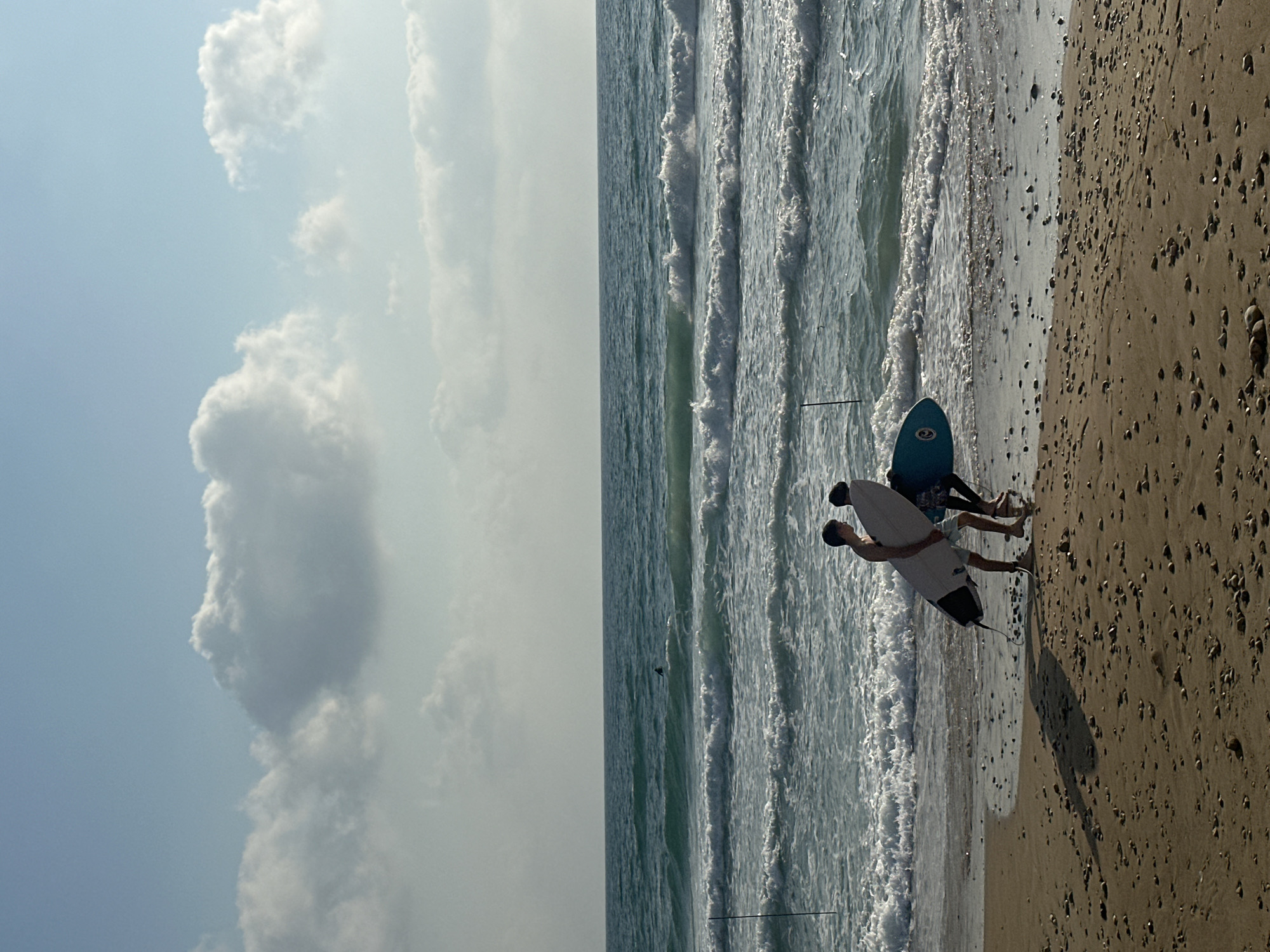 Two individuals, likely young men, are walking on a beach in Haifa, Israel, carrying surfboards. The individual on the left is carrying a white surfboard under his right arm. The individual on the right is carrying a blue surfboard with a circular emblem. Both are walking away from the water and towards the foreground. The beach is sandy with scattered pebbles. The ocean is visible in the background, with waves breaking near the shore. The sky is partly cloudy with a light blue hue. A lone surfer is visible further out in the ocean. The scene appears to be in the late afternoon or early evening, given the angle of the shadows cast by the individuals.